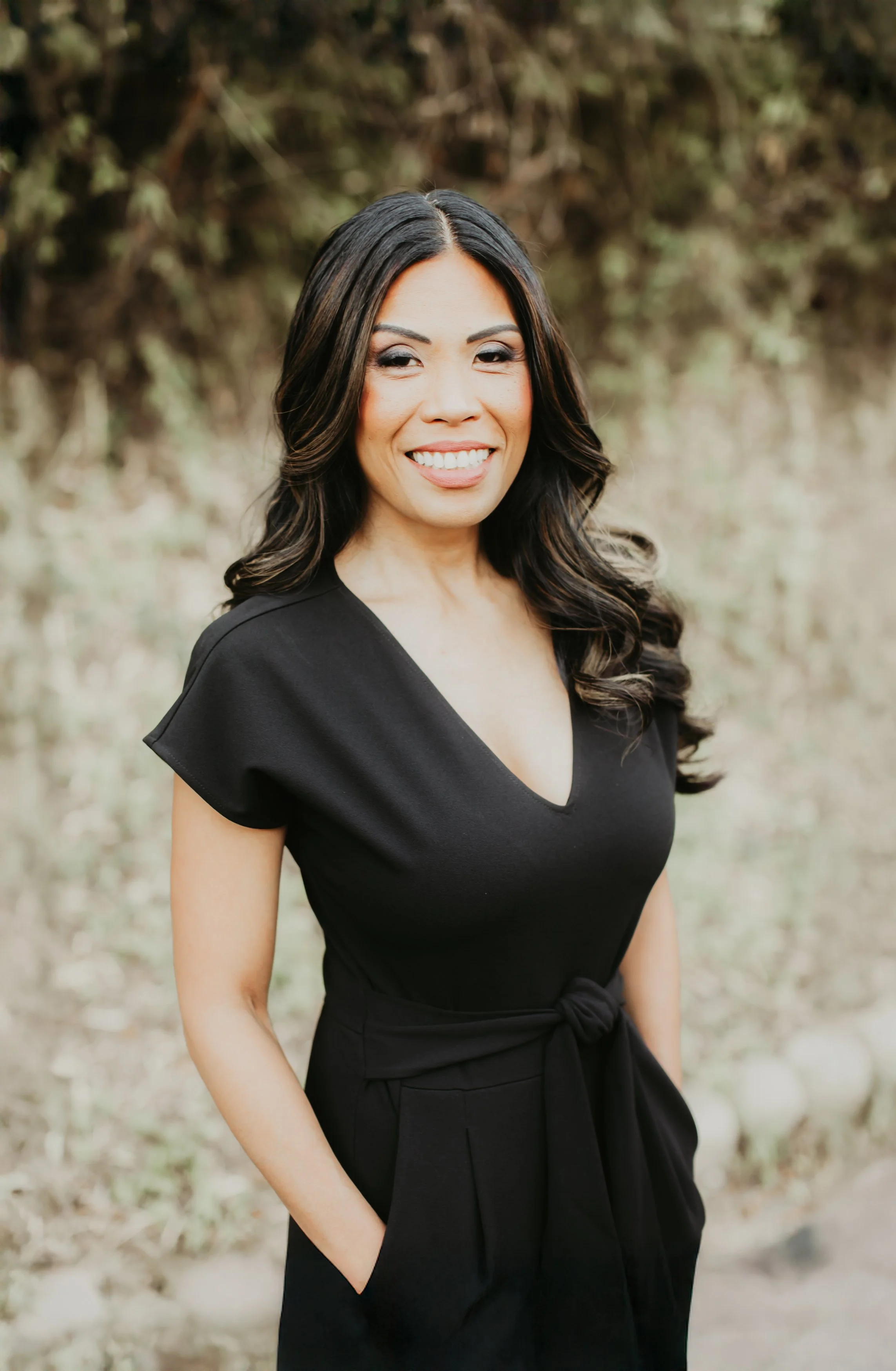 Portrait of a woman with long, wavy dark hair, smiling, wearing a black dress, standing outdoors with a blurred natural background.