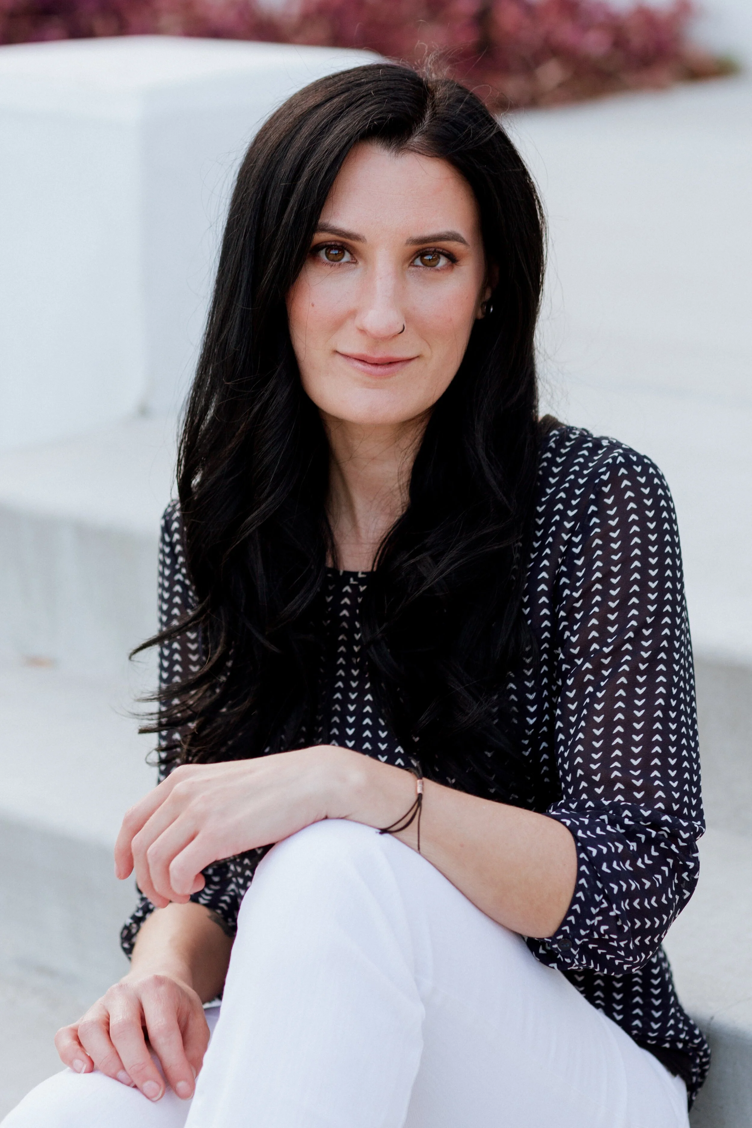 A woman with long black hair, brown eyes, and light skin sitting outdoors, wearing a black and white patterned top and white pants. She is looking at the camera with a slight smile, with a blurred background of pink foliage.