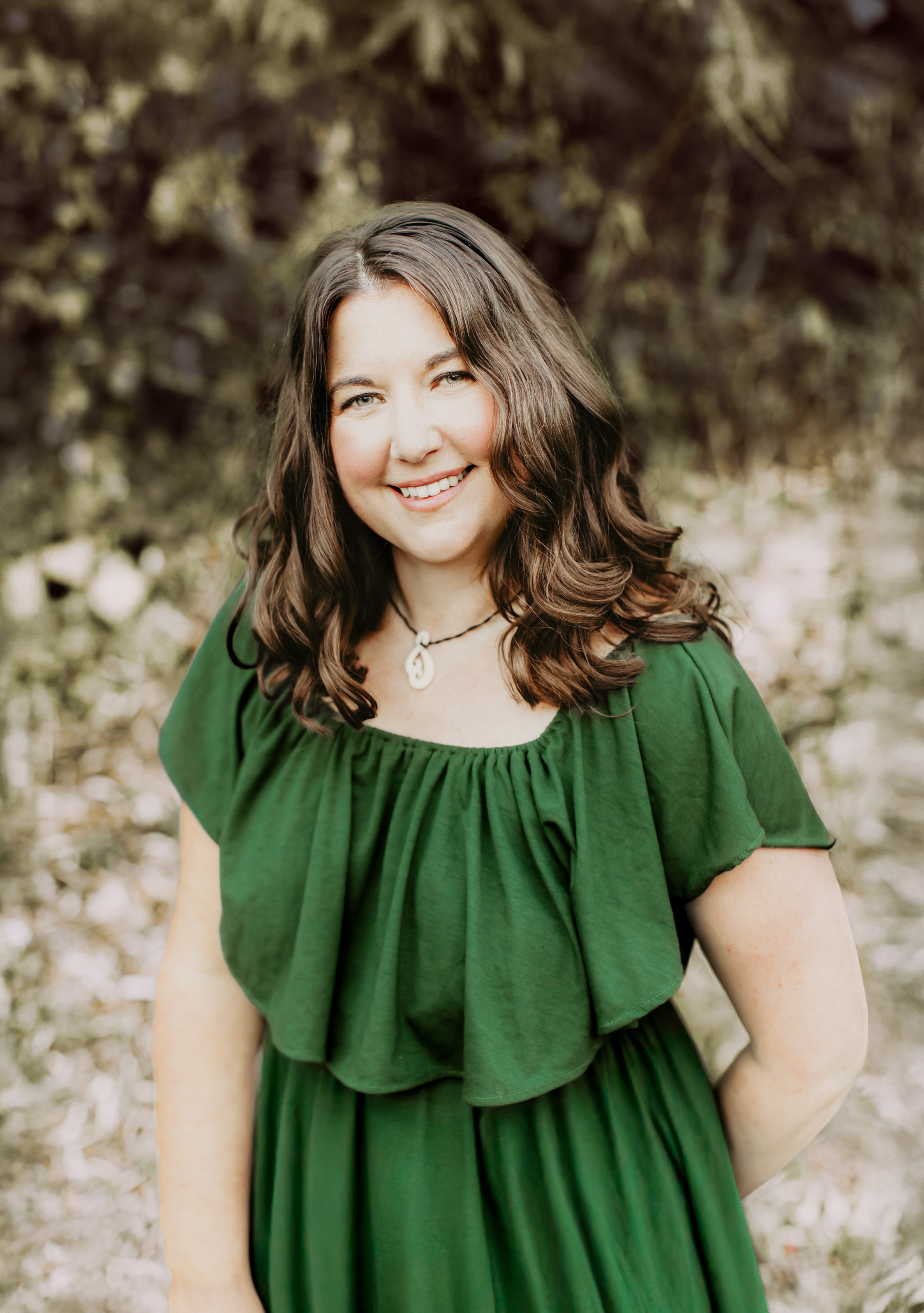 A smiling woman with brown wavy hair, wearing a green dress and a shell necklace, standing outdoors with blurred trees and foliage in the background.
