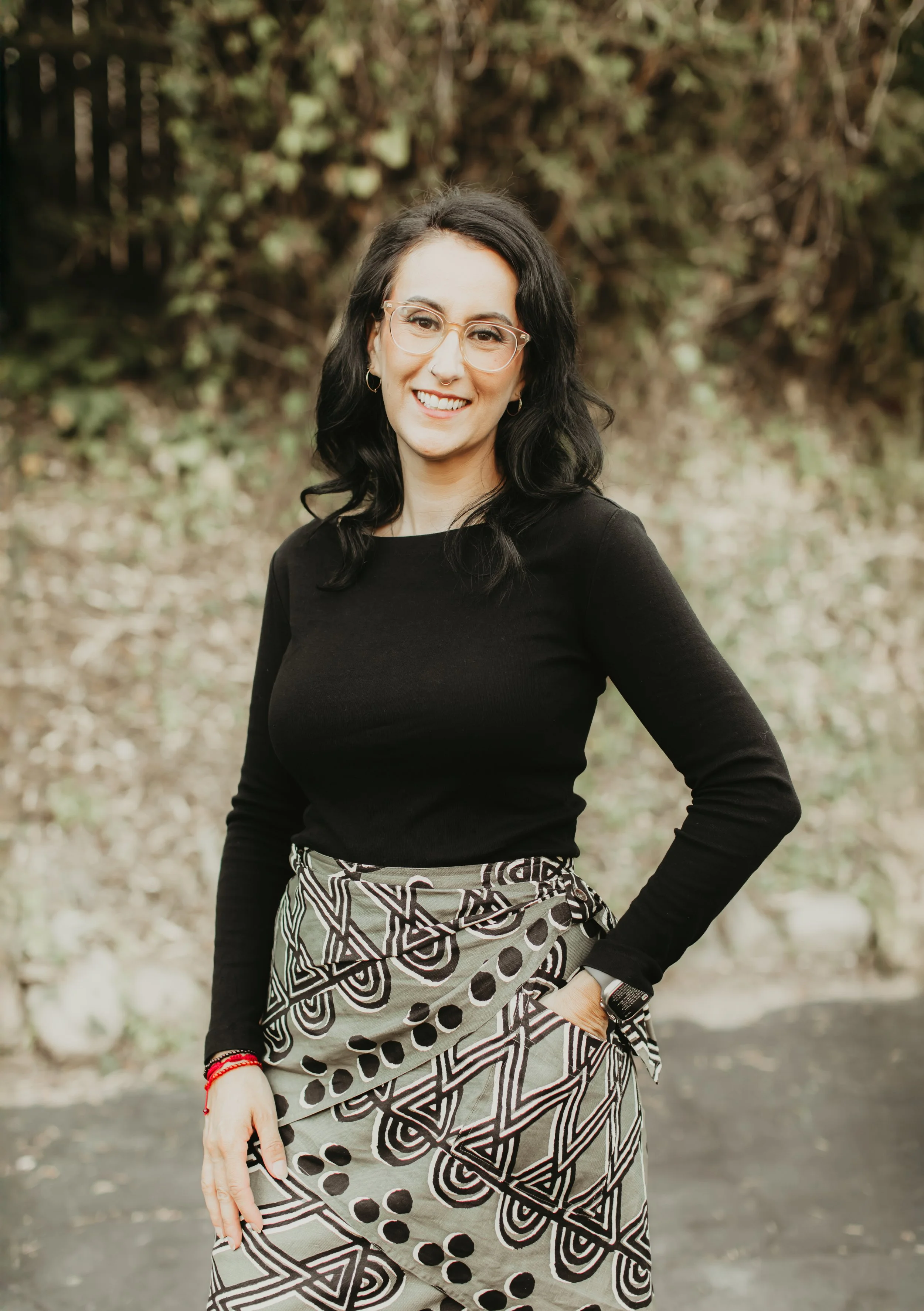 A woman with black hair, glasses, and hoop earrings standing outdoors with a blurred background of trees and bushes, wearing a black top and a patterned skirt.