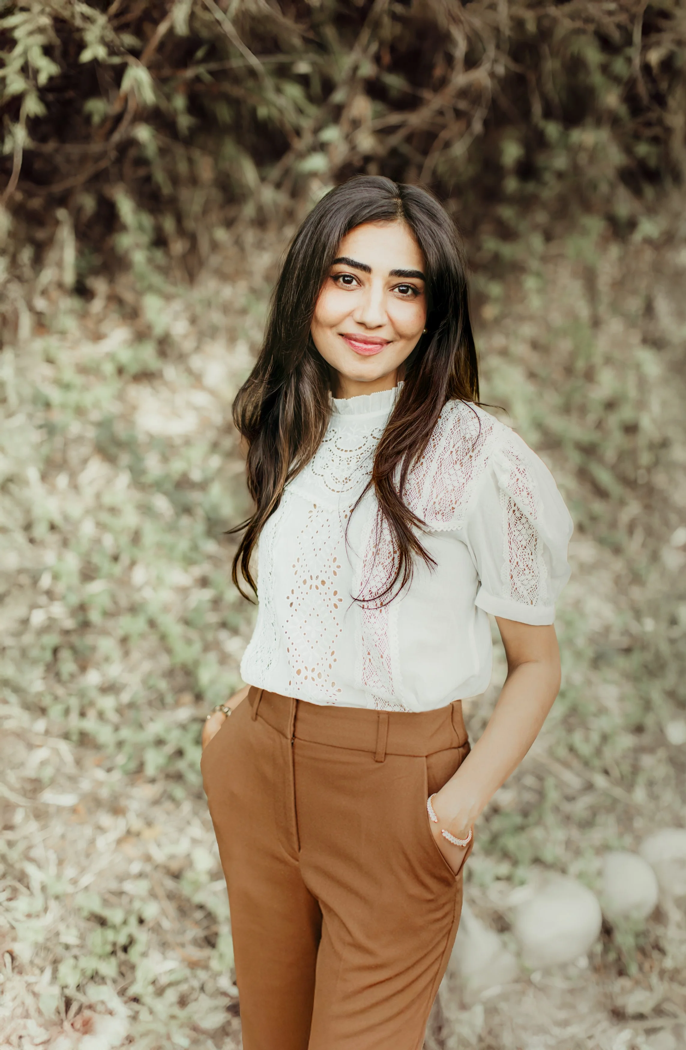A young woman with long dark hair smiling, wearing a white embroidered blouse and brown trousers, standing outdoors with a blurred background of trees and foliage.