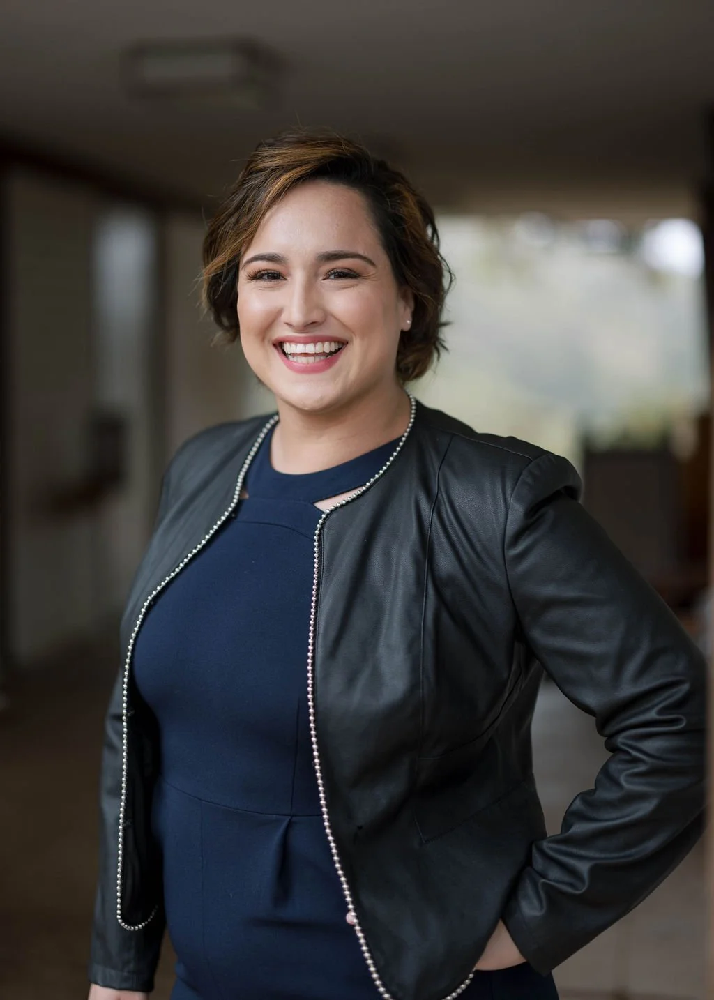 A young woman with short brown hair, smiling, wearing a black leather jacket and a navy blue top, standing indoors with a blurred background.