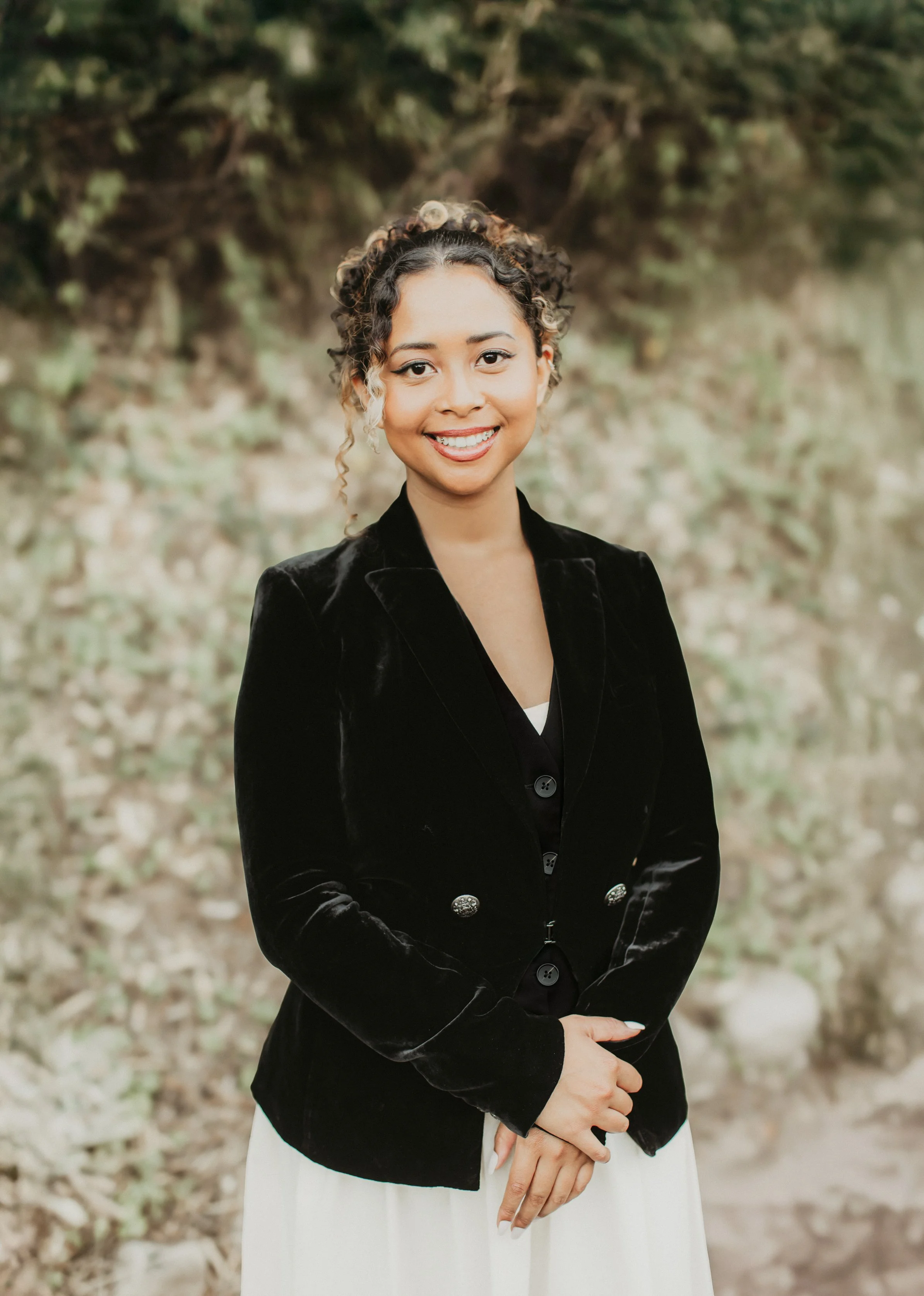A woman smiling outdoors, with dark curly hair and wearing a black velvet jacket and a white skirt, standing in front of a blurred green and brown natural background.