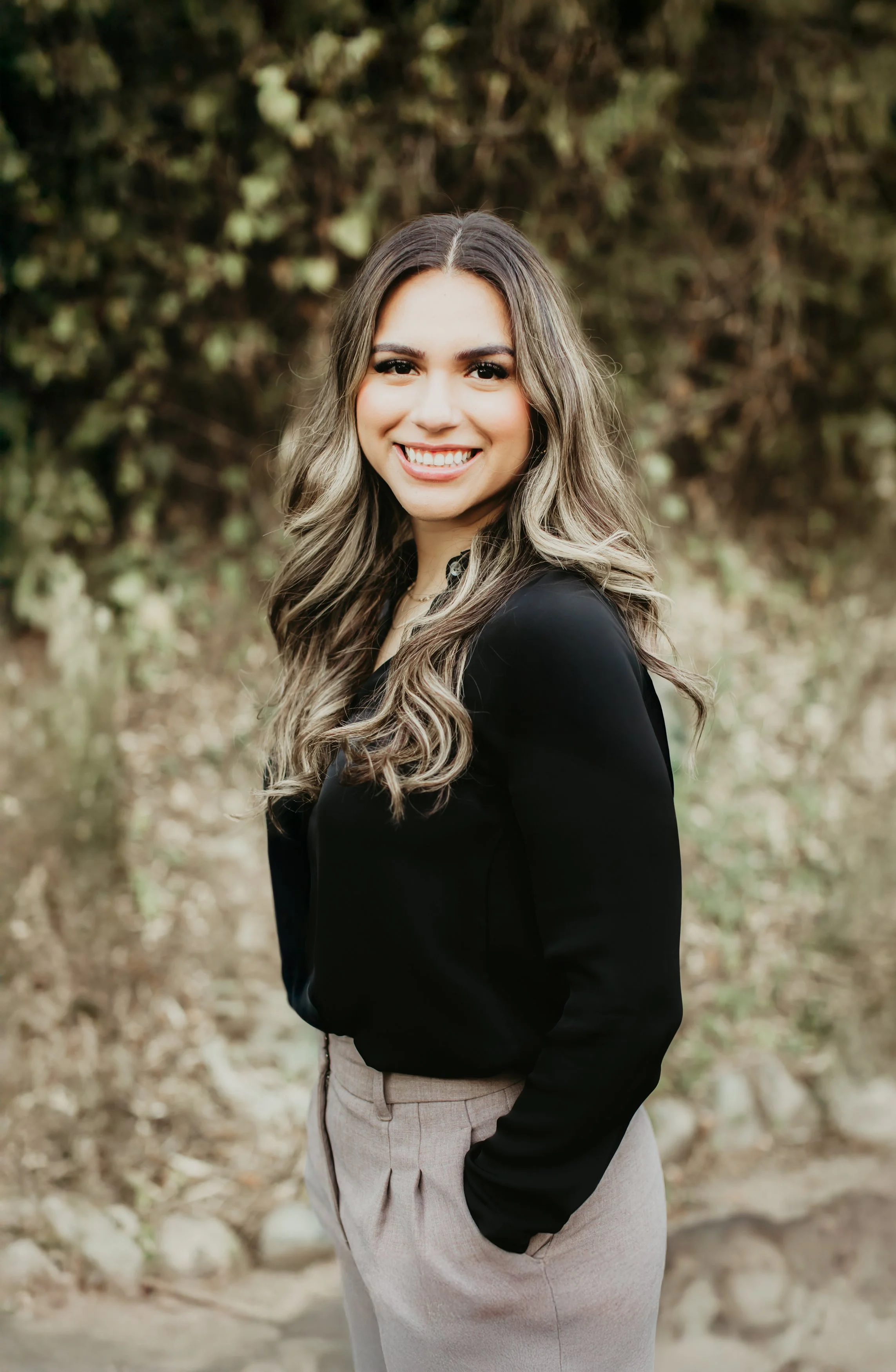 A young woman with long wavy hair, smiling, wearing a black top and beige pants, standing outdoors with a natural background of trees and rocks.