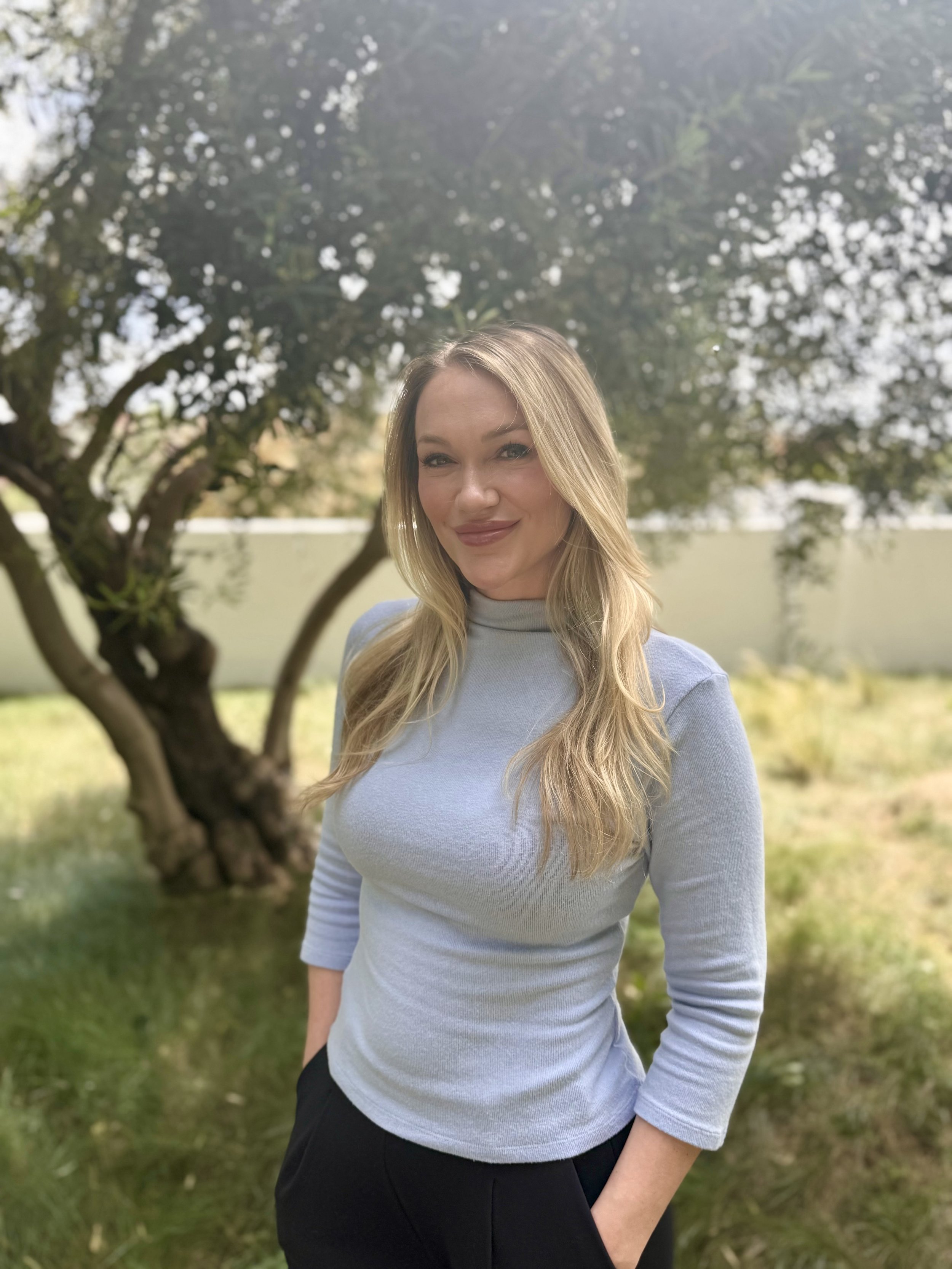 A woman with long blonde hair wearing a light gray long-sleeved shirt and black pants standing outdoors near a tree with green grass and foliage in the background.