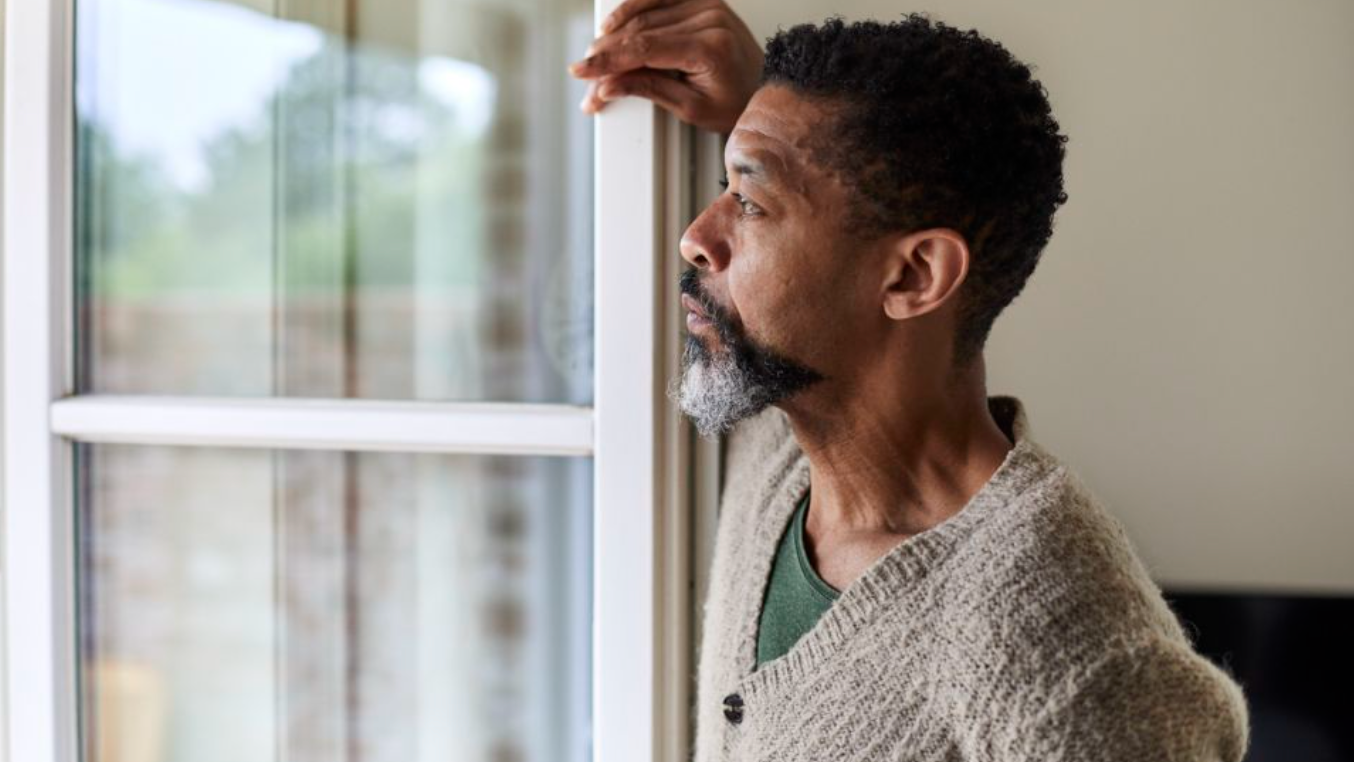 A man with a beard and curly hair looking thoughtfully outside through a window, resting his hand on the window frame.