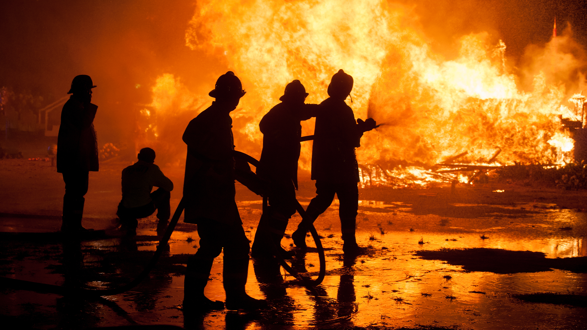 Firefighters silhouetted against a large blaze at night, spraying water on flames.