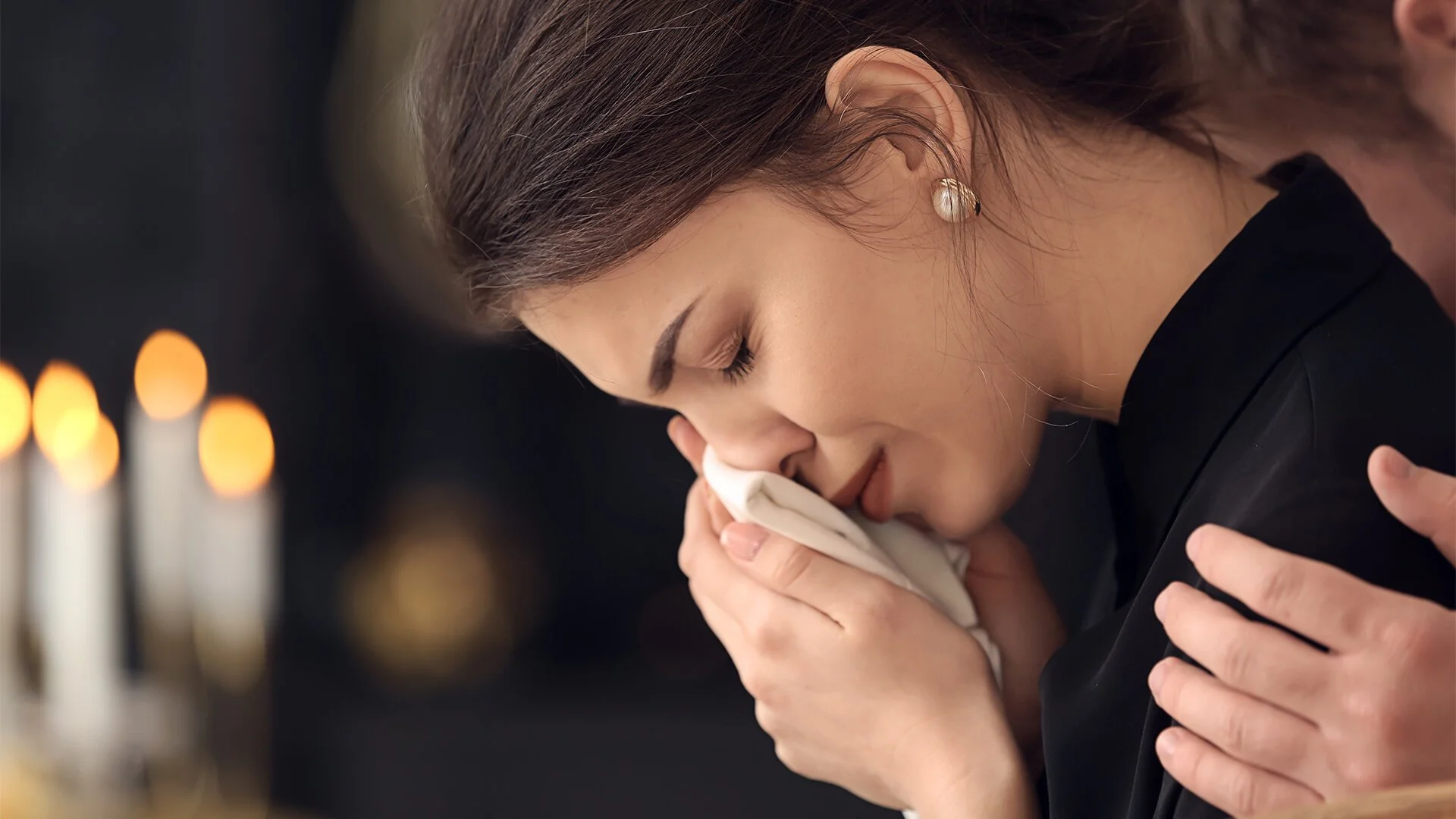 A woman with closed eyes, wearing pearl earrings, is holding a white cloth near her face, possibly praying or mourning. In the background, blurred candles are visible.