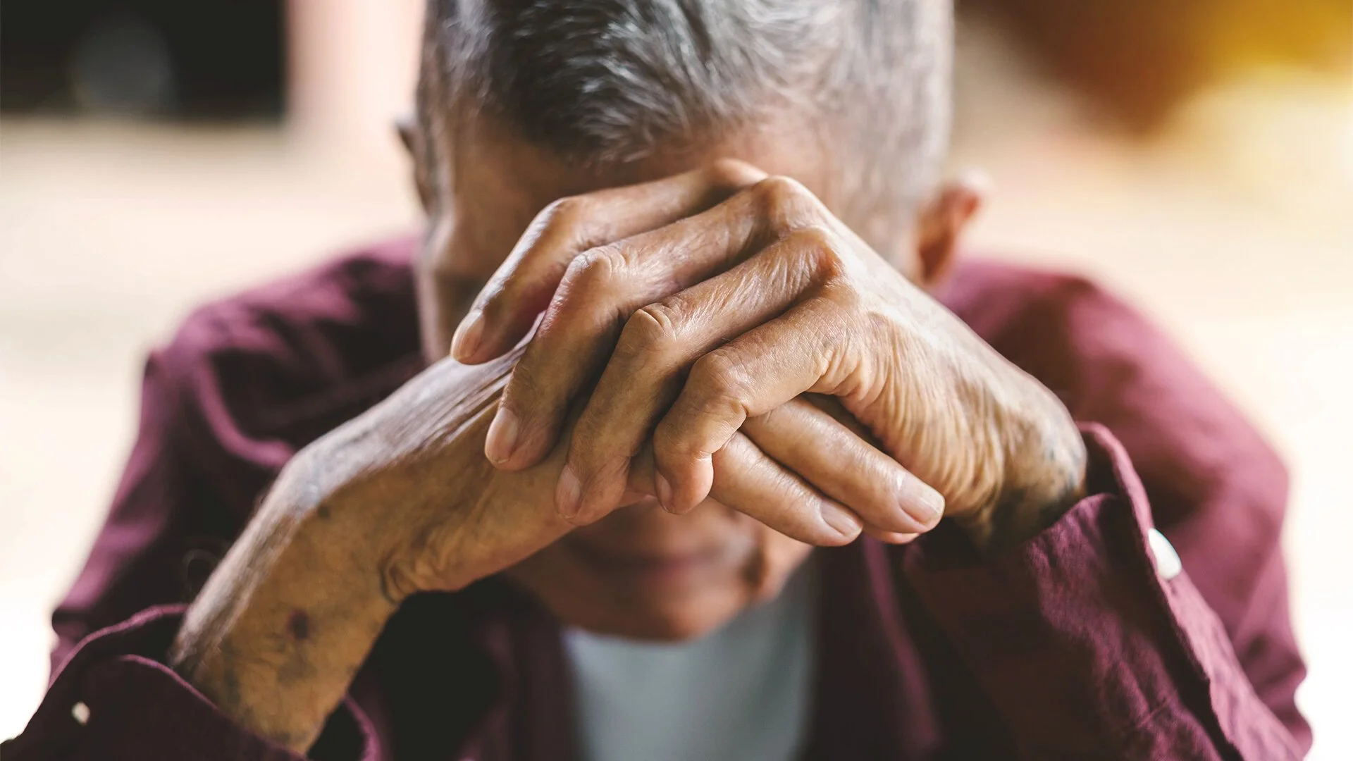 An elderly person with gray hair and aged hands, wearing a maroon shirt, sits with their hands clasped over their face, showing signs of aging and emotional distress.