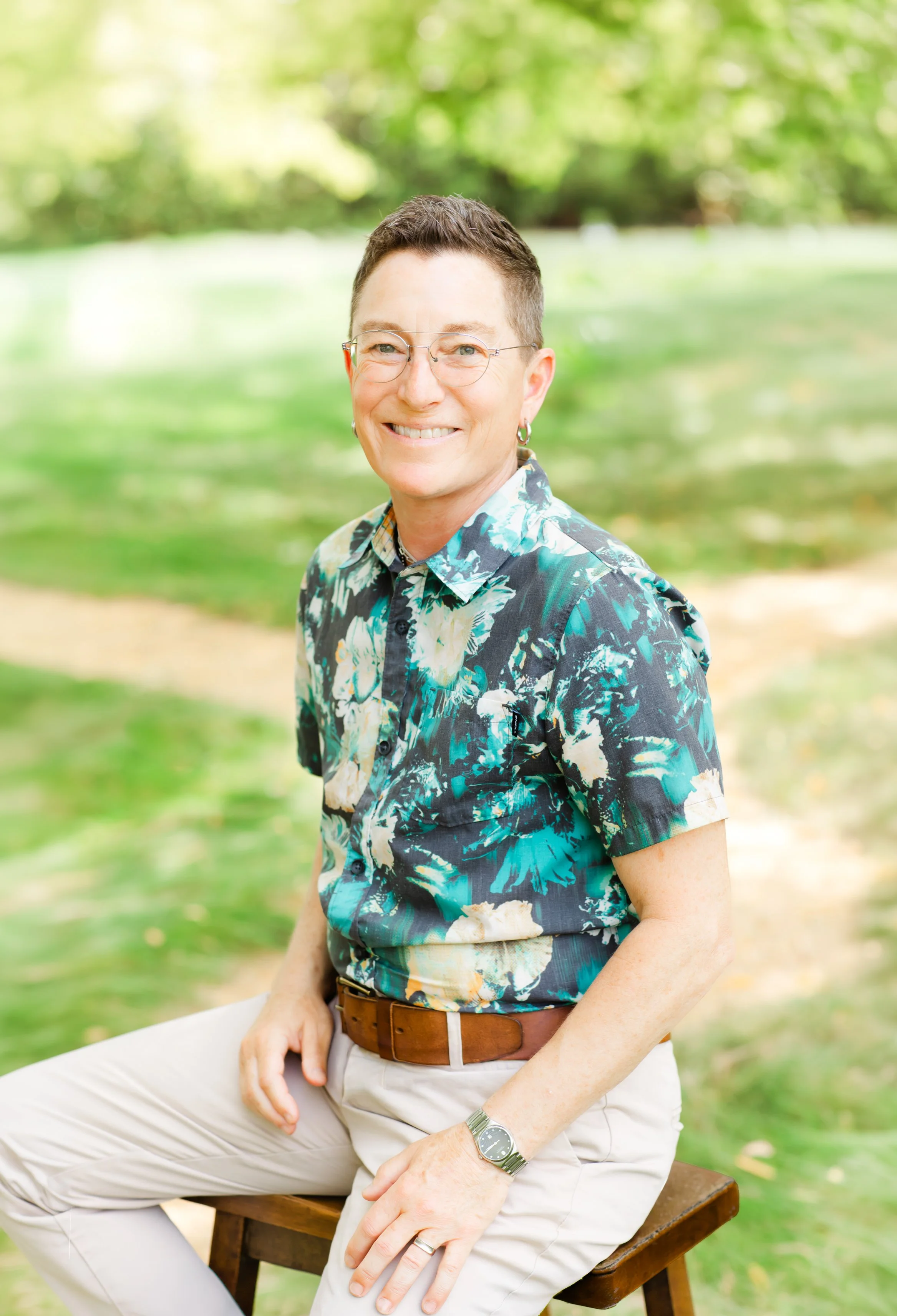 A woman with short hair and glasses, wearing a blue blazer and a patterned shirt, sitting outdoors with a blurred background of trees with yellow and green leaves, smiling at the camera.