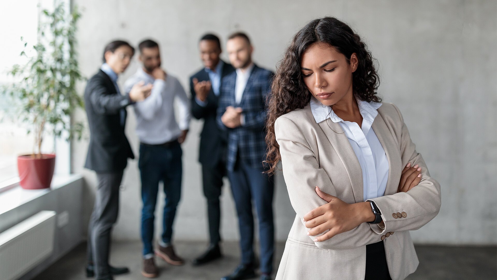A woman in a beige blazer with arms crossed and head down, appears upset, while a group of four colleagues in business attire stand in the background, engaged with their phones and conversations, in an office setting.