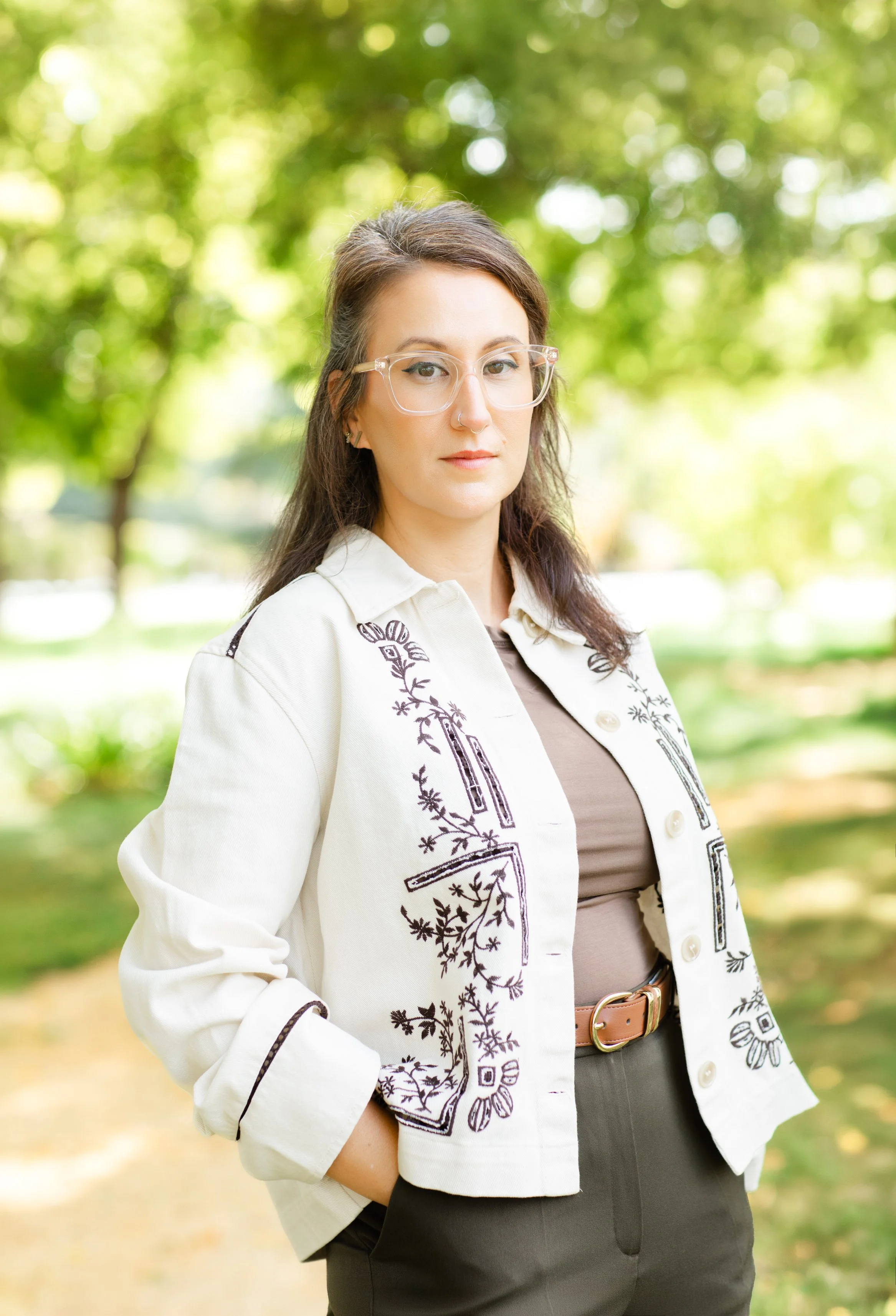 A woman with black hair, glasses, and hoop earrings standing outdoors with a blurred background of trees and bushes, wearing a black top and a patterned skirt.