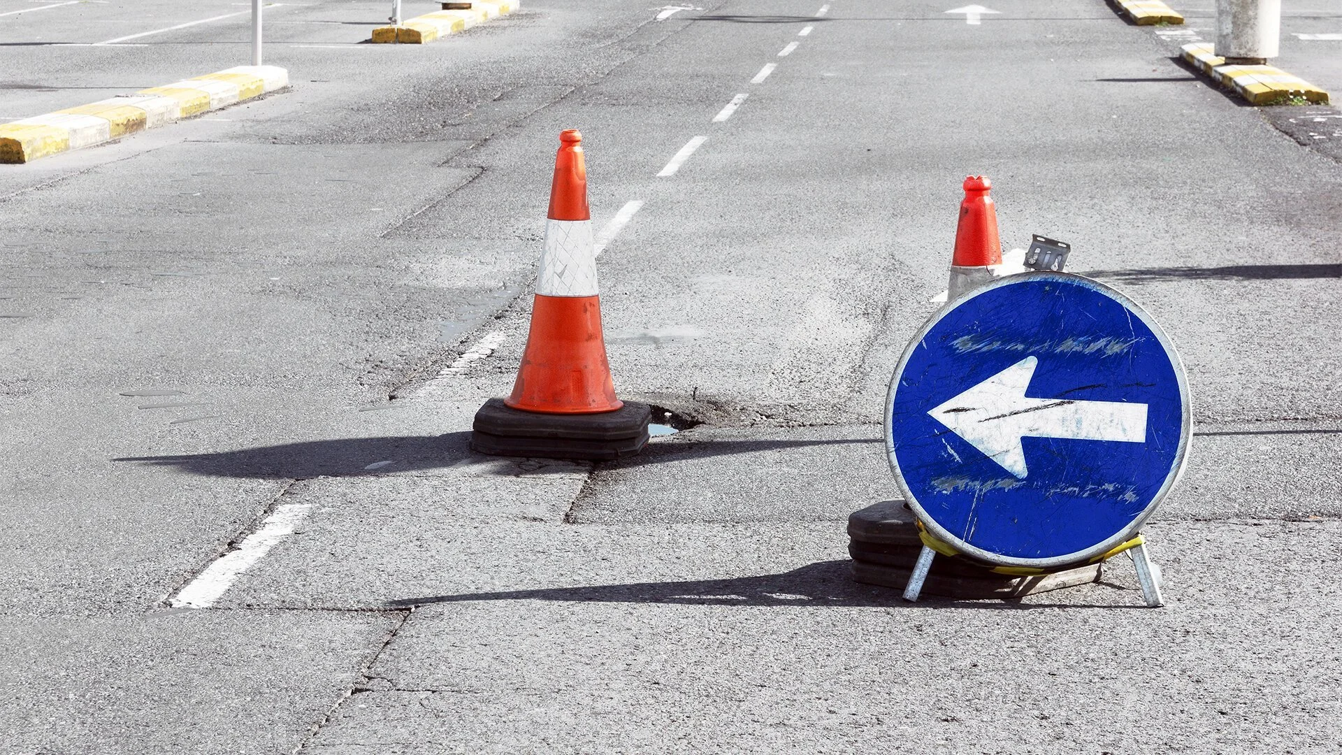 Orange traffic cone and a circular blue road sign with a white left arrow, both placed on a road with traffic barriers and dashed lane markings.