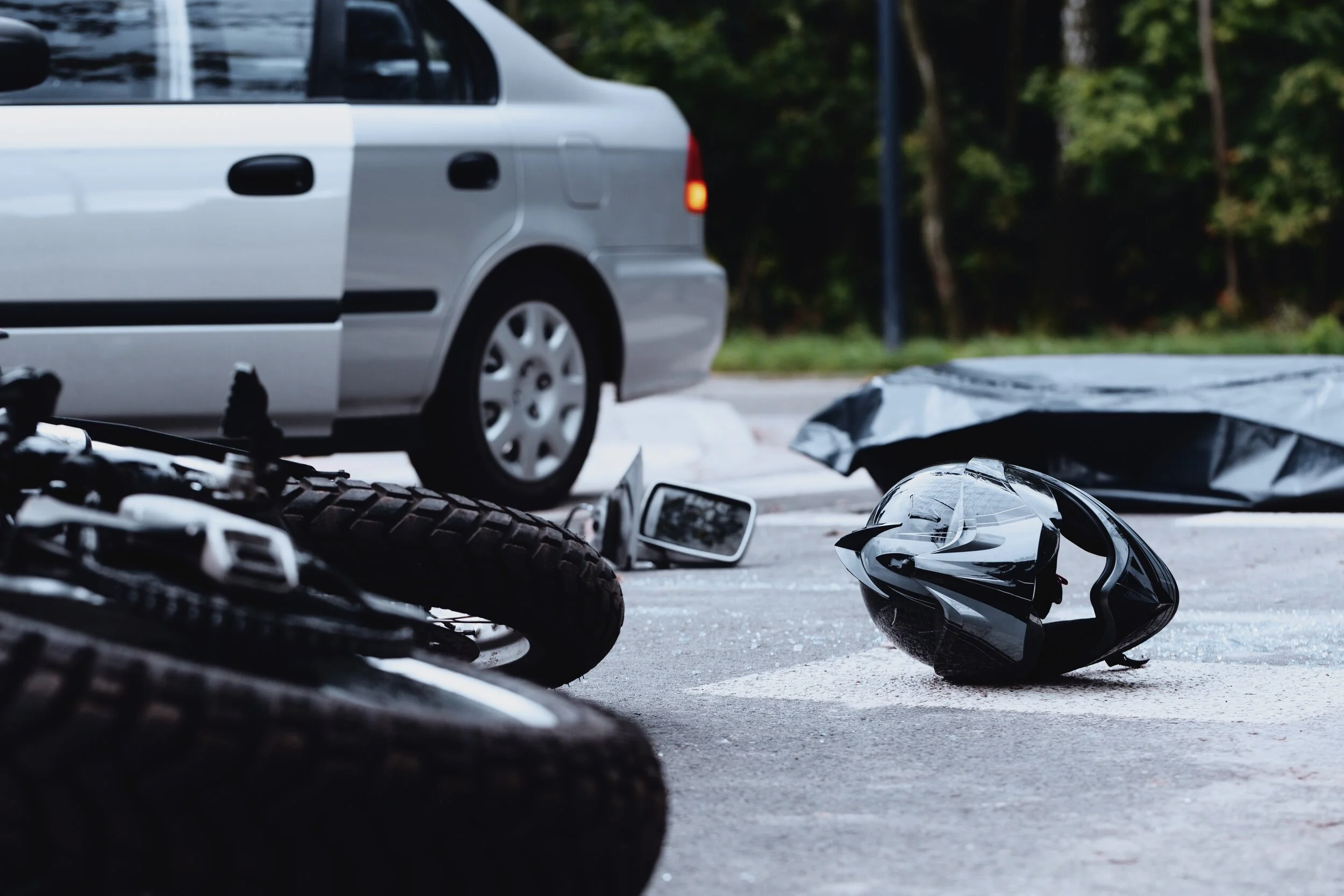Scattered motorcycle helmets and a motorcycle on the ground in the foreground, a silver car stopped on a street, and trees in the background.