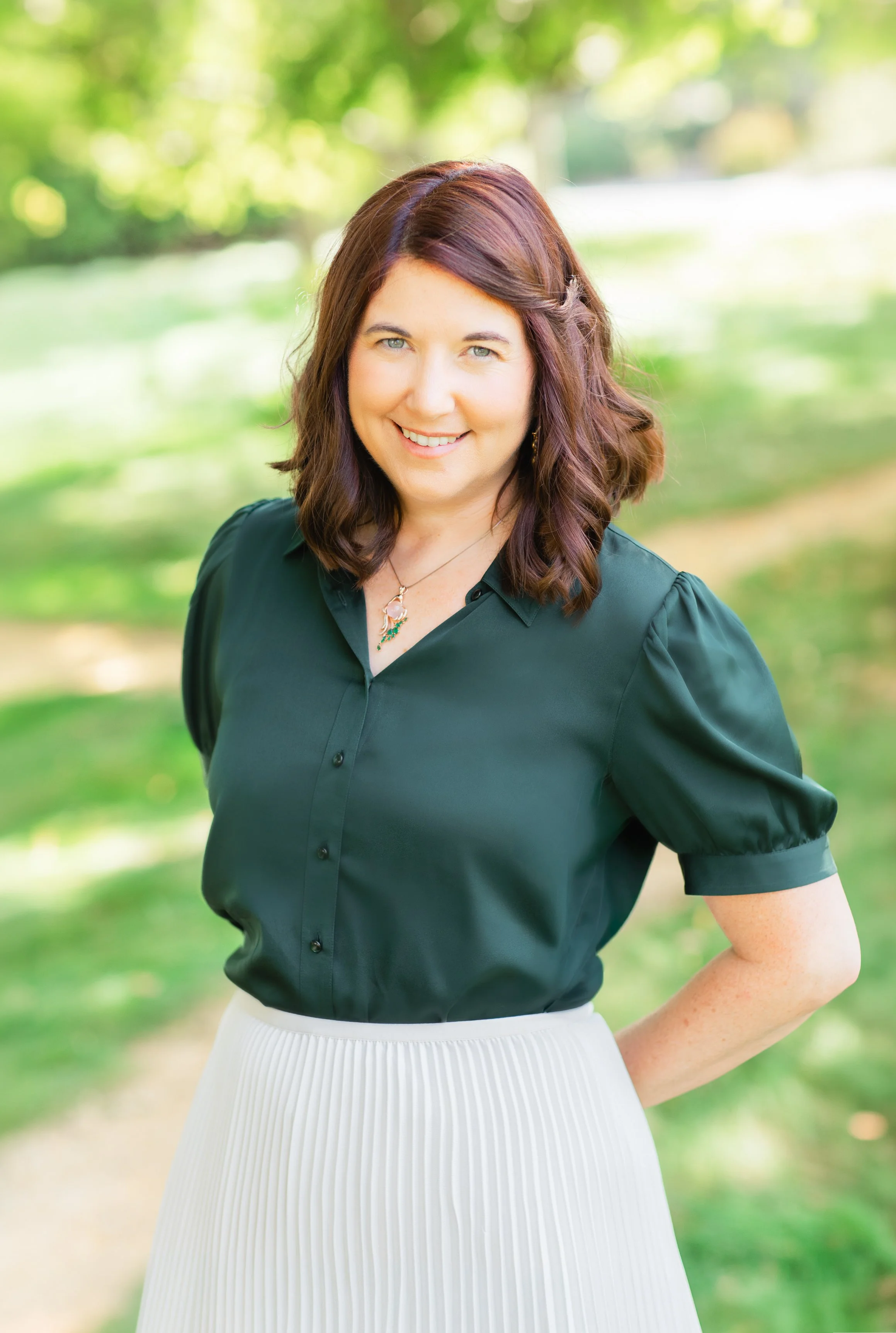 A smiling woman with brown wavy hair, wearing a green dress and a shell necklace, standing outdoors with blurred trees and foliage in the background.
