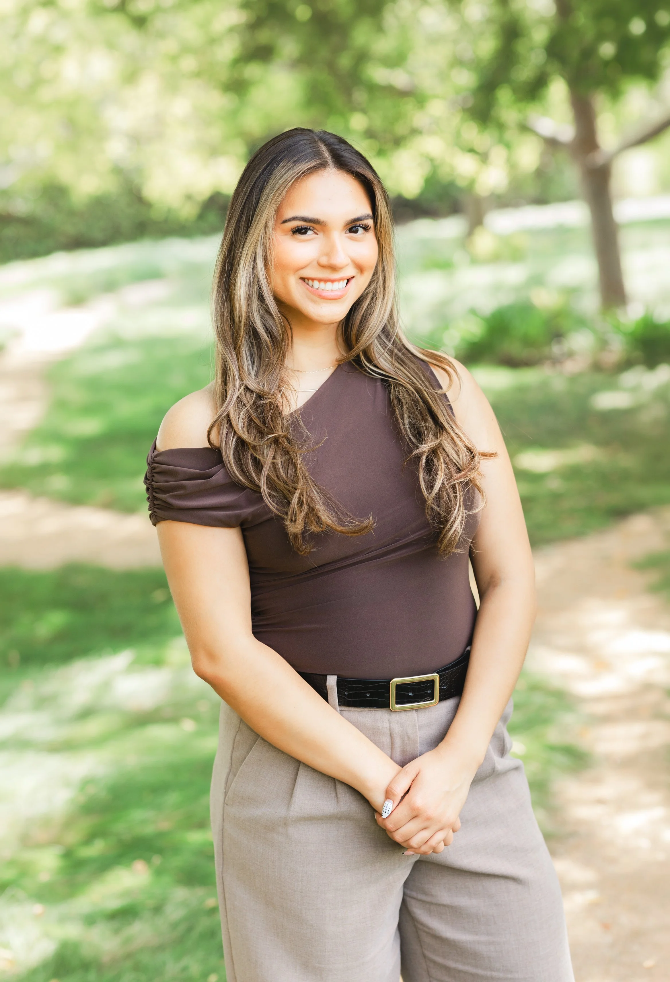 A young woman with long wavy hair, smiling, wearing a black top and beige pants, standing outdoors with a natural background of trees and rocks.