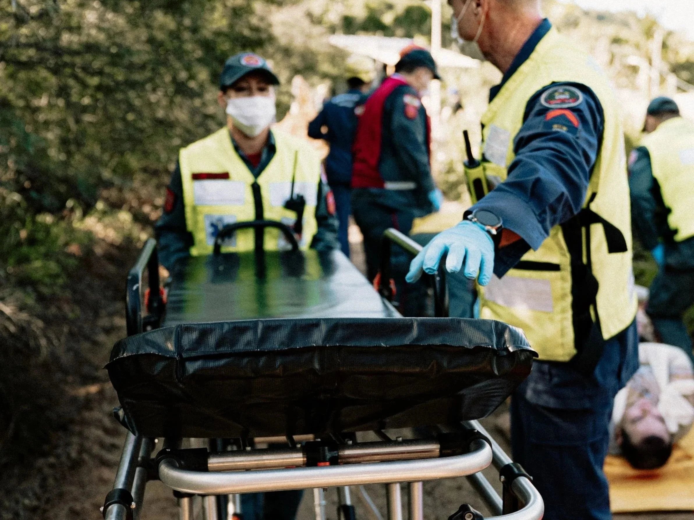 People wearing yellow safety vests and masks standing outdoors, with one person pointing at a wheelchair or cart in front of them.