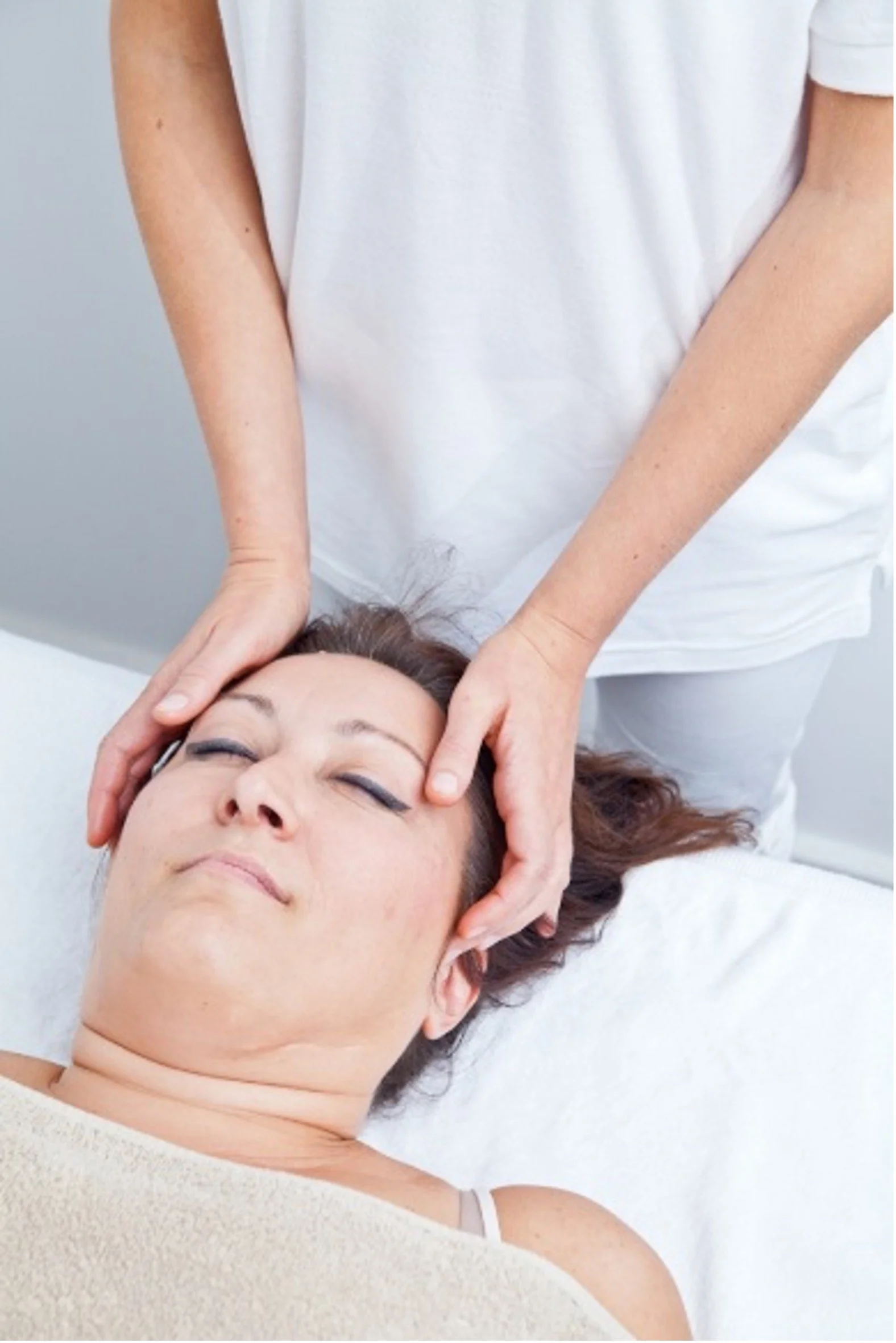 The face & shoulders of a woman lying on her back on a massage couch, looking relaxed with  eyes closed, with a professional therapist standing behind, lightly resting their hands on either side of her face, with a beige towel covering her upper body