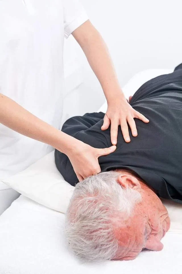 Hands of a healthcare practitioner performing Bowen Therapy moves on the shoulder of an elderly man, who is laying face down on a massage couch, wearing a dark shirt.