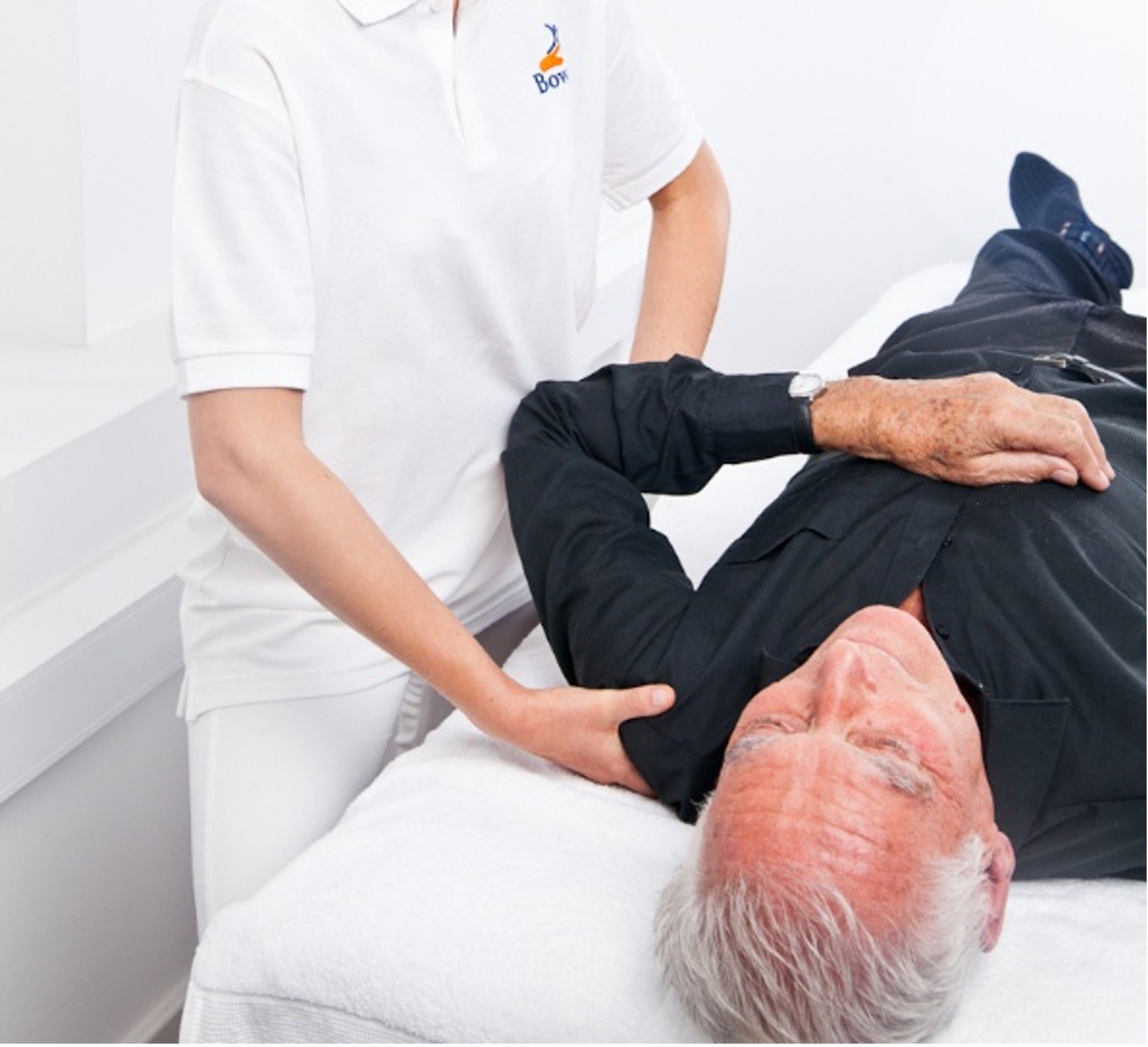 A practitioner in a white top bearing the logo of the Bowen Therapy Professional Association, standing next to a massage couch giving Bowen treatment on shoulder & arm of an elderly man lying on his back with eyes closed, wearing  a dark shirt.