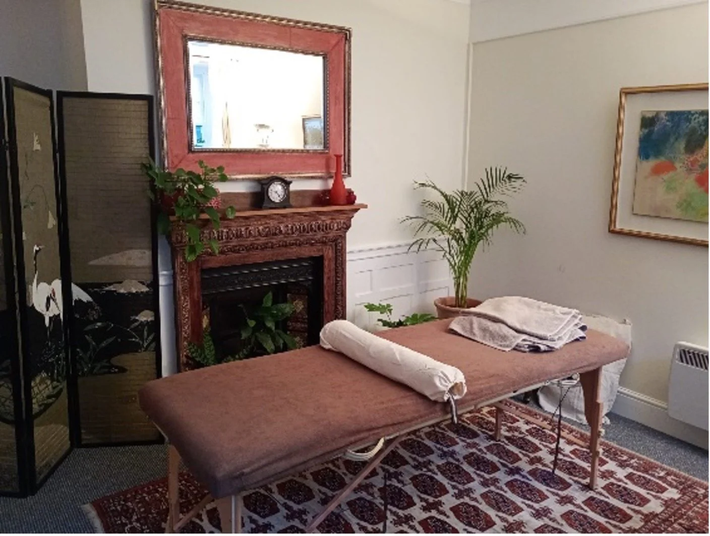 Massage couch with a brown cover, bolster & folded towels on top, in a room with a decorative fireplace, a mirror, potted plants, framed artwork, and a folding privacy screen.
