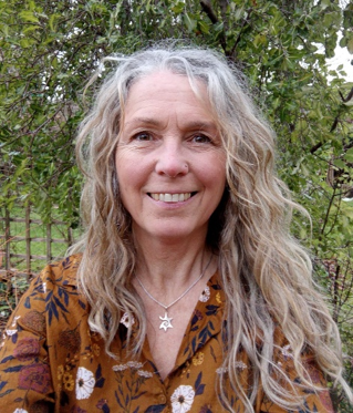 A smiling woman with long, wavy, light hair, outdoors with green bushes in the background.