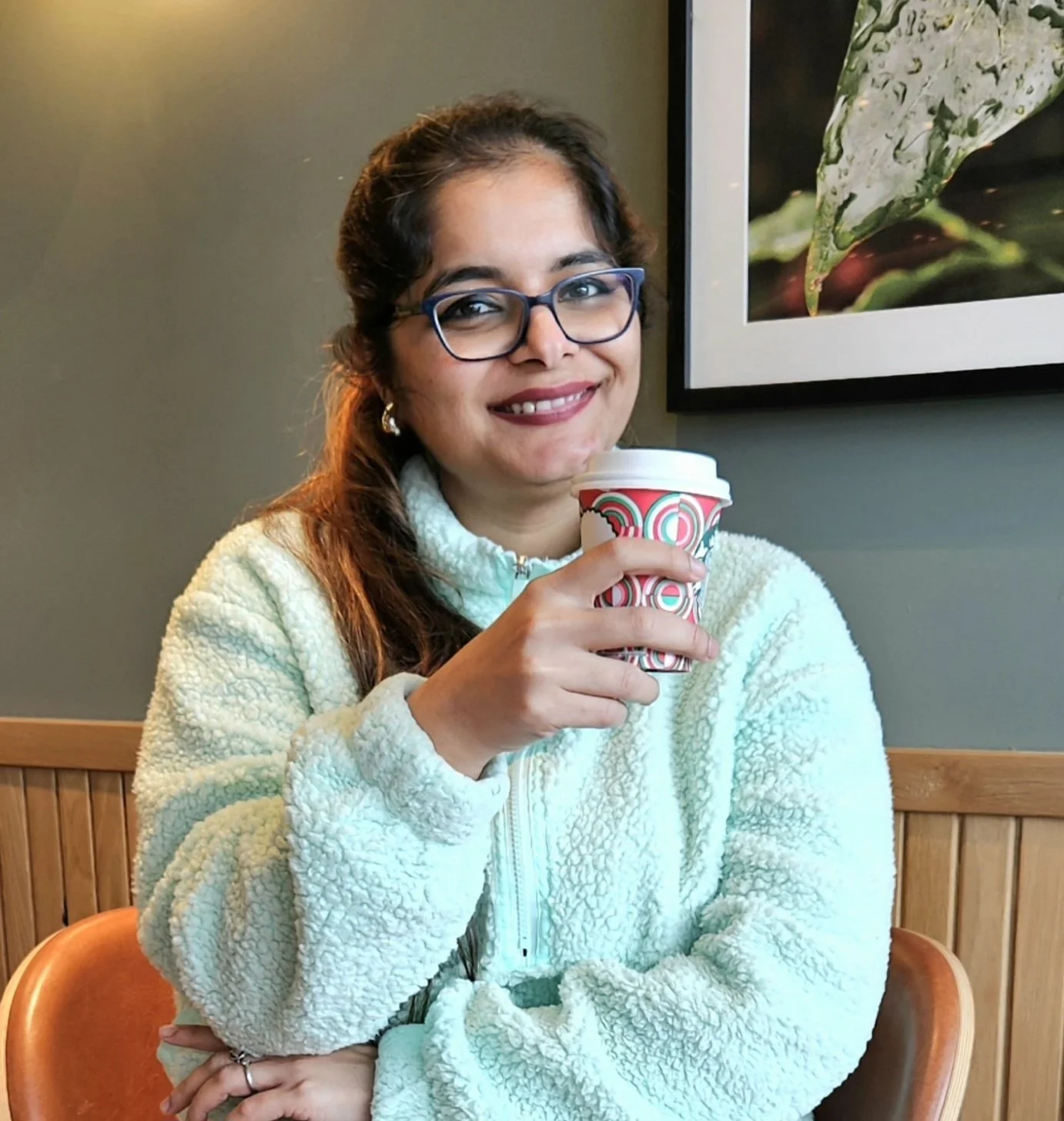 A woman with glasses and earrings, smiling, holding a colorful cup and sitting on a wooden chair inside a café, near a framed nature photograph on the wall.