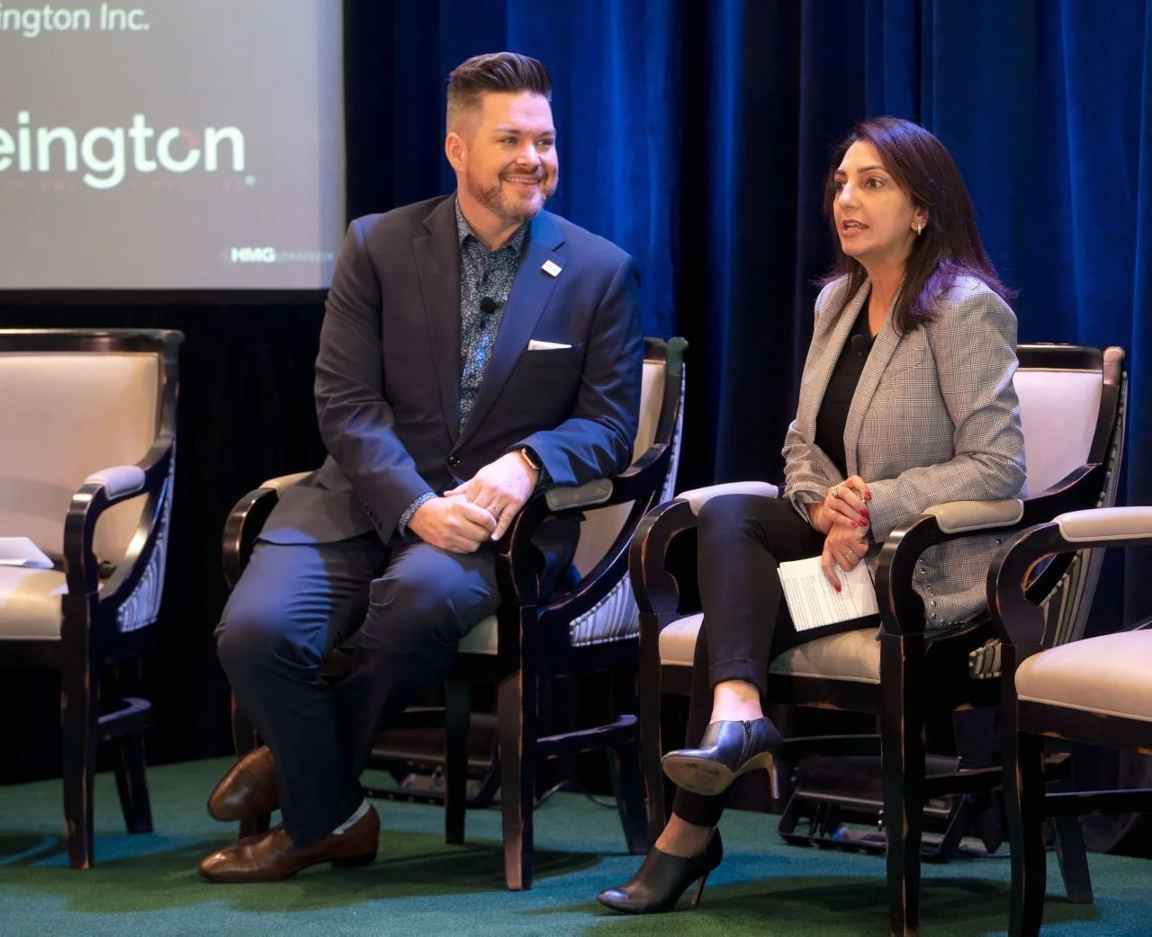 Two people sitting and conversing on a stage during a panel discussion or conference. The man has short, styled hair and a beard, wearing a dark suit with a patterned shirt, and the woman has shoulder-length dark hair, wearing a checkered blazer, bla