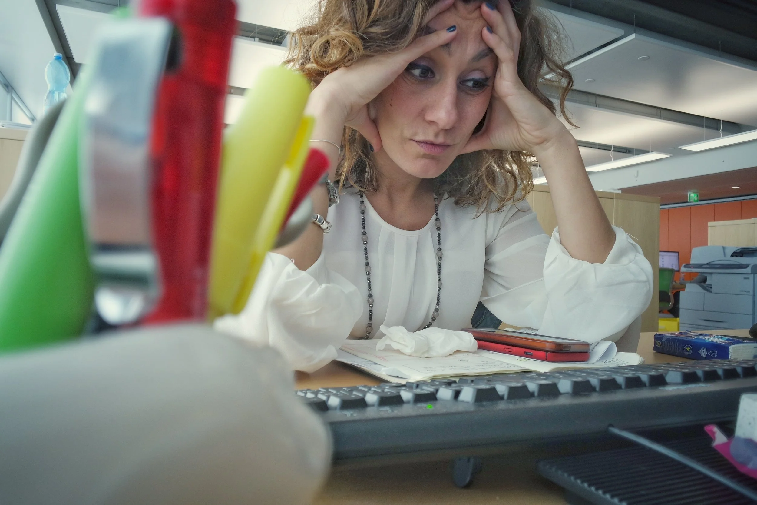 A woman with curly hair and a white top sitting at a desk, holding her forehead with both hands, appearing stressed or overwhelmed in an office environment.