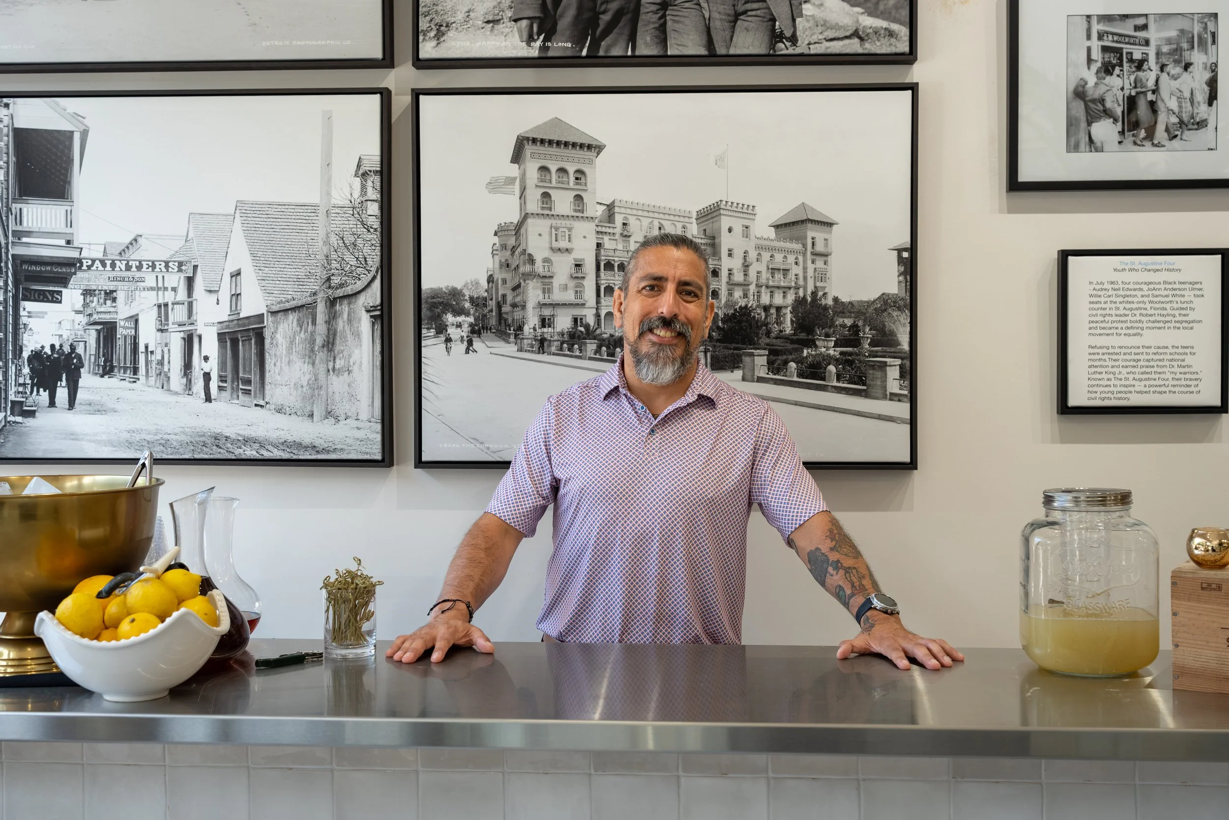 A smiling man with a beard and tattoos on his arm, wearing a checkered shirt, stands behind a kitchen counter in front of a wall decorated with black and white framed photos of City scenes and buildings.