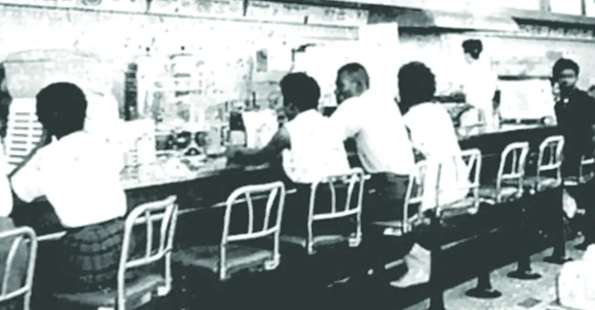Black and white photo of a bar with six men seated at the counter, some drinking and some talking.