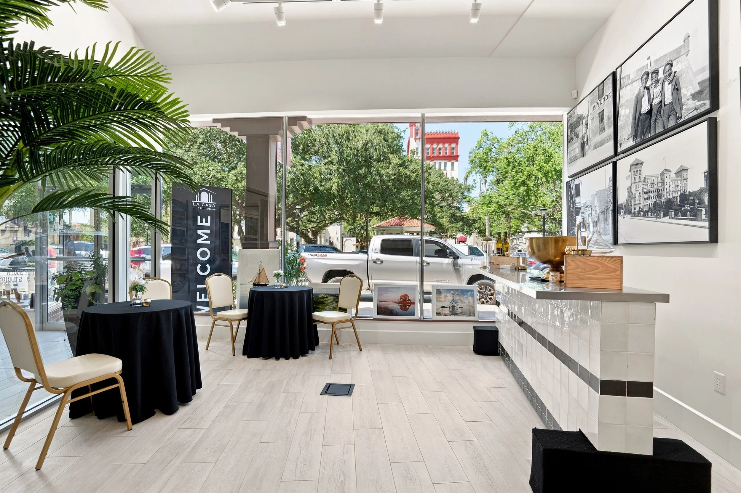 Interior view of a modern art gallery or showroom with large windows, potted plants, round tables with white chairs, and black tablecloths. Black and white photographs of buildings and people are displayed on the walls. There is a reception or display counter with decorative items, and a large green plant in the foreground.