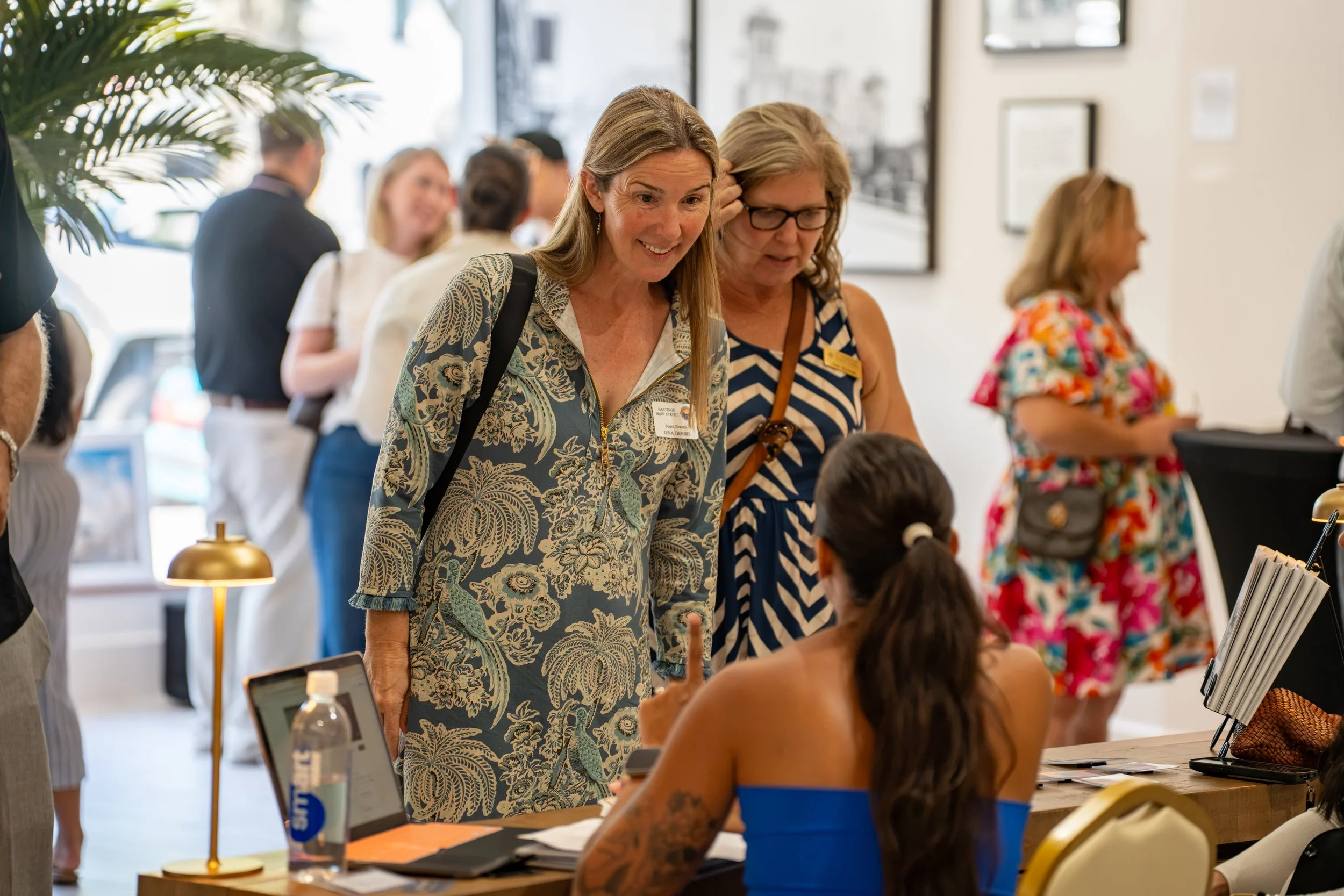 People engaging at a registration or check-in desk during an event or conference, with women interacting and using laptops and tablets.