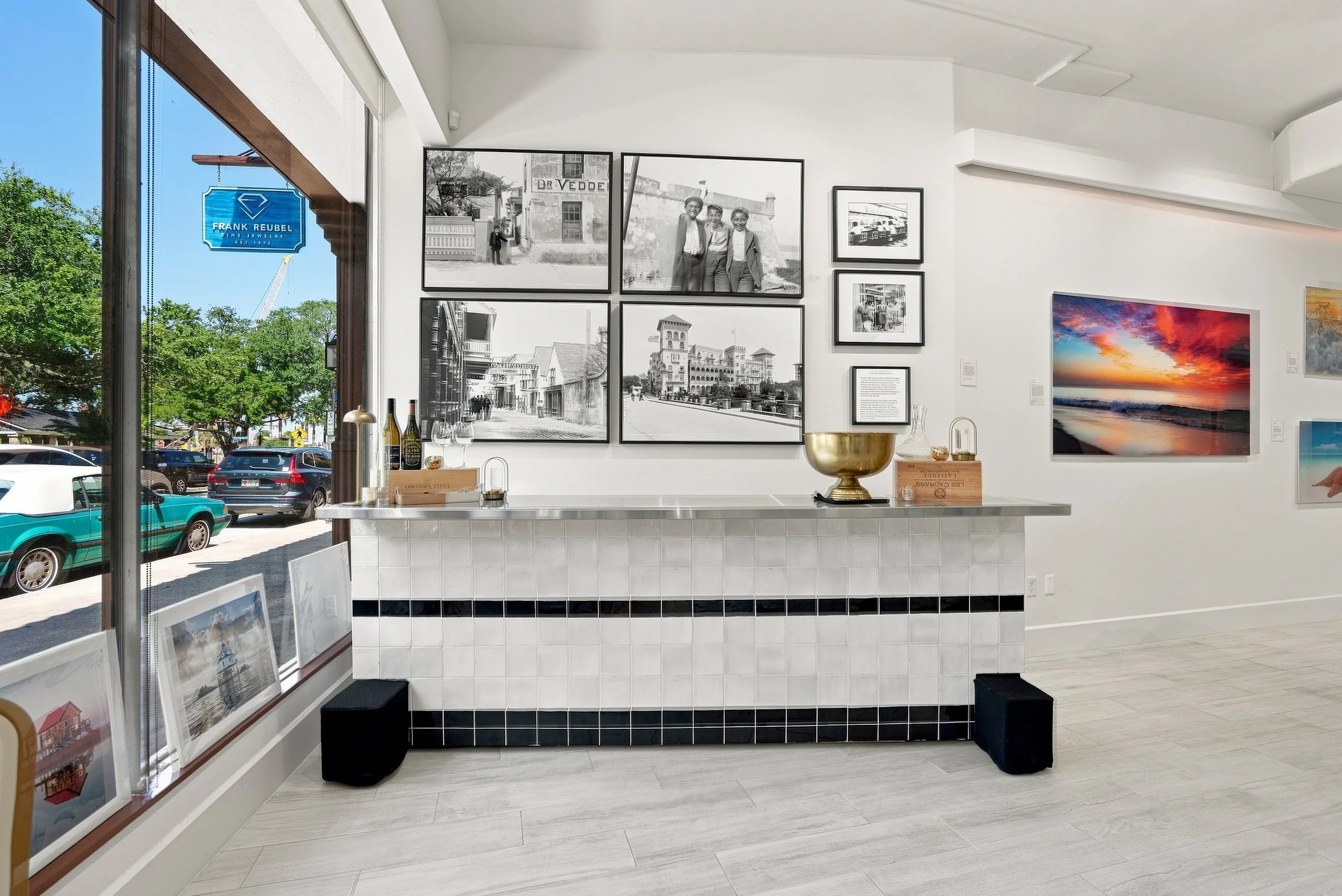 An art gallery interior with black and white photographs on the wall, colorful sunset photograph, framed artwork leaning against the window, a display table with bottles and glasses, and two small black stools. Sunlight streams through large glass windows revealing parked cars outside.