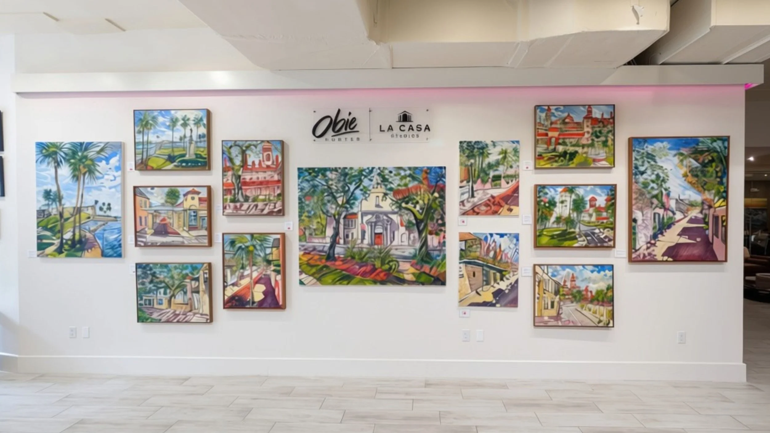 An art gallery room with white walls and ceiling, featuring multiple framed landscape photographs. The room has modern lighting, a green sofa with pink and purple flowers on a wooden table, and brown leather chairs. There are plants and decorative items displayed around the room.