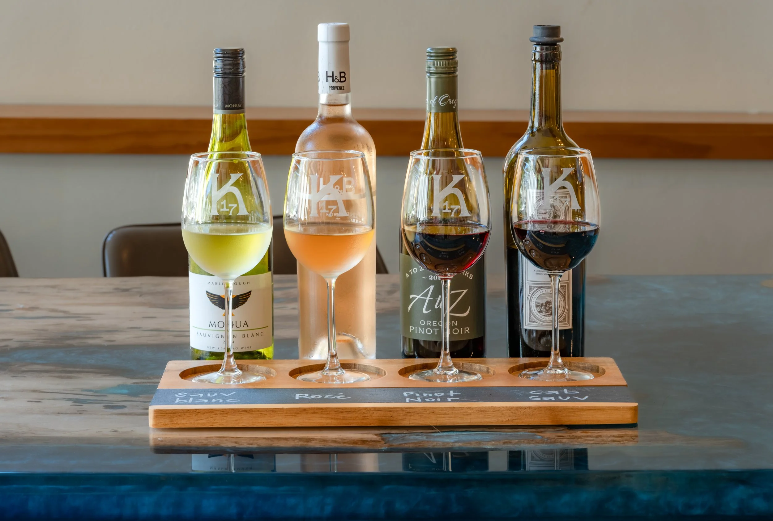 Four wine bottles and four wine glasses filled with different types of wine are arranged on a wooden tasting board on a marble table. The background is a neutral wall with a wooden trim.