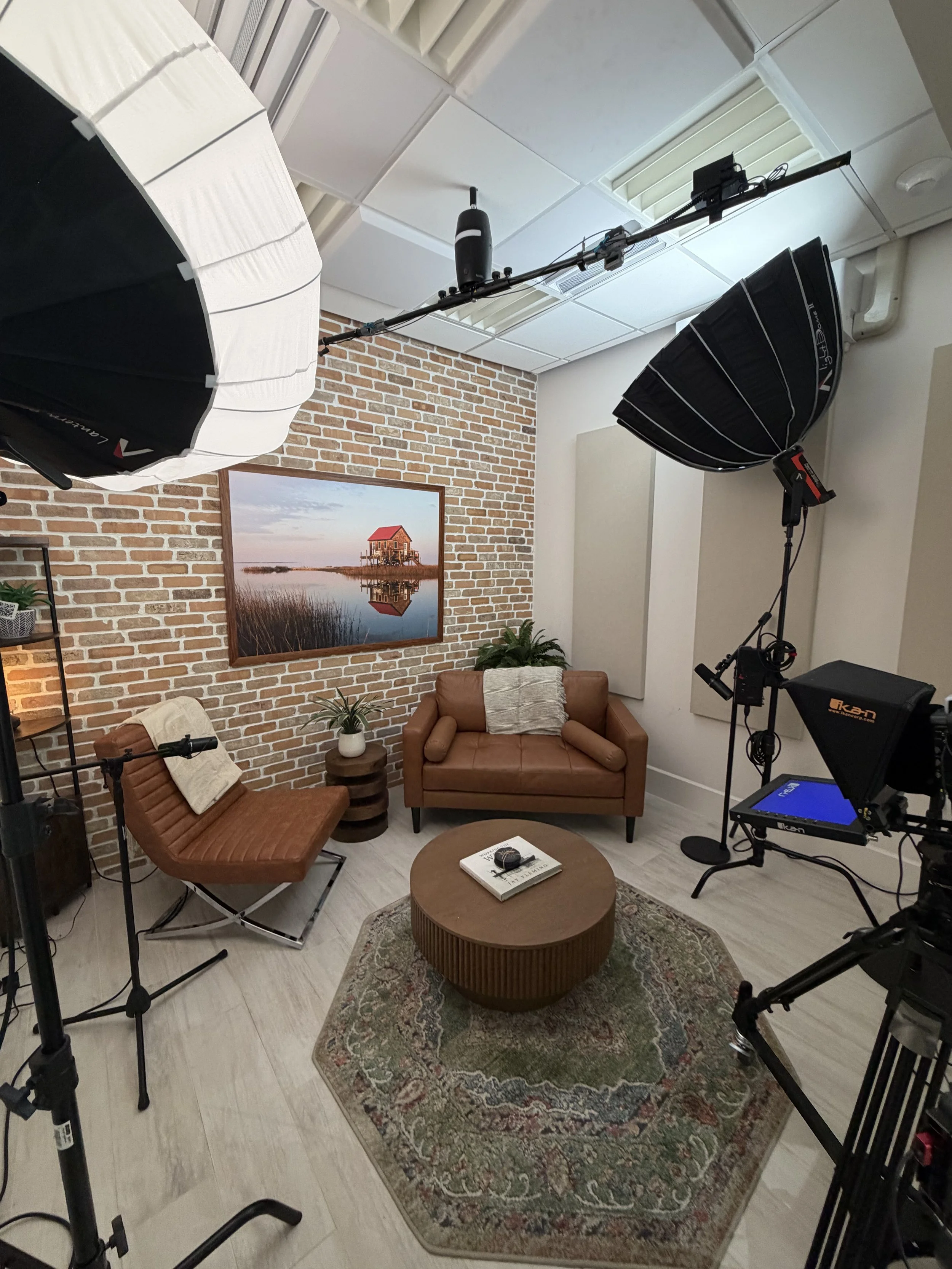An indoor studio setup with professional lighting and camera equipment. Includes a brick wall with a framed landscape photograph of a house near water, two brown chairs, a small round coffee table with a book and headphones, some plants, and acoustic panels on the walls.