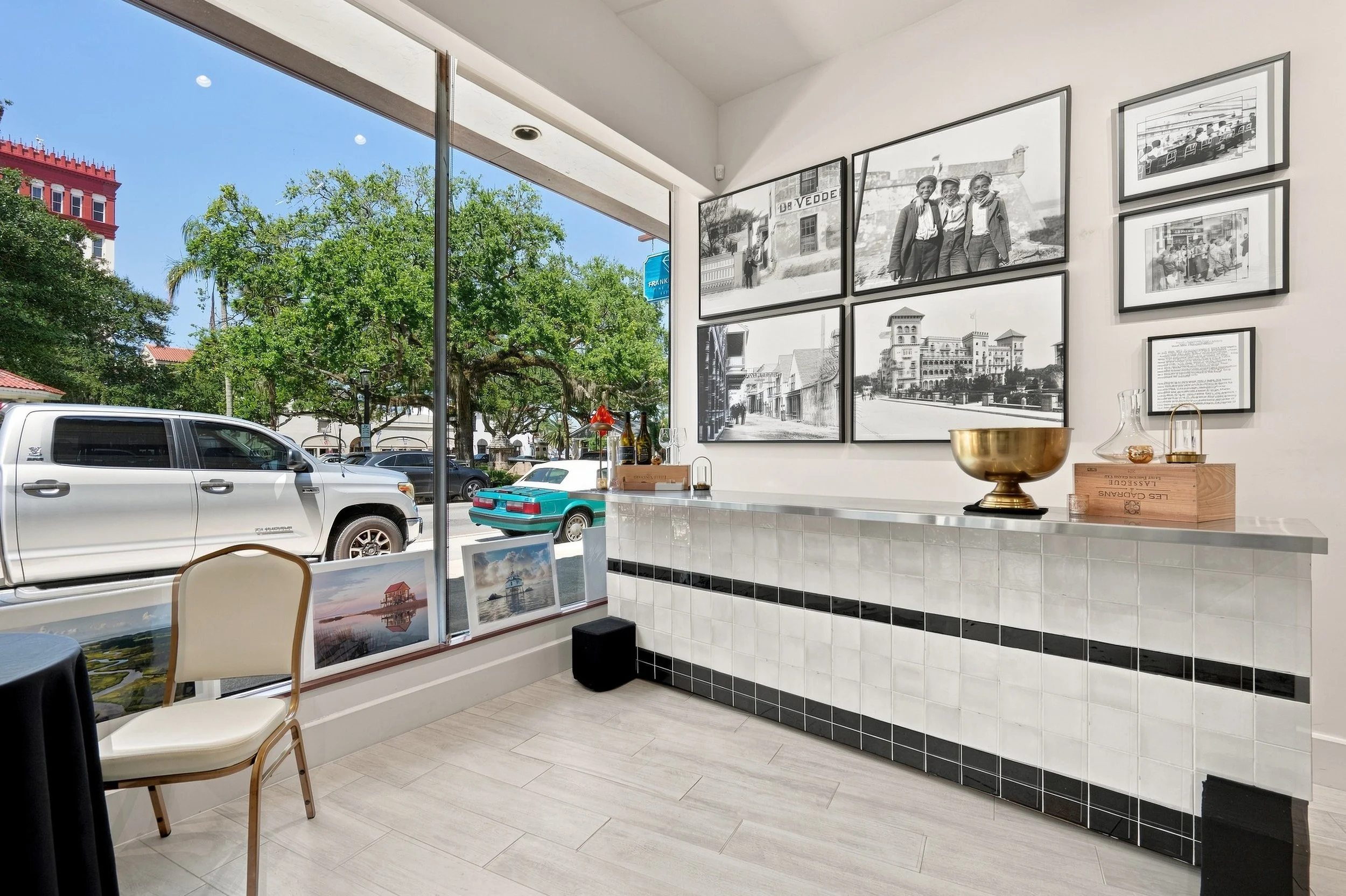 Interior of a gallery or cafe with black and white framed photographs on the wall, a countertop with decorative items, and a large window showing parked cars, trees, and a blue sky outside.