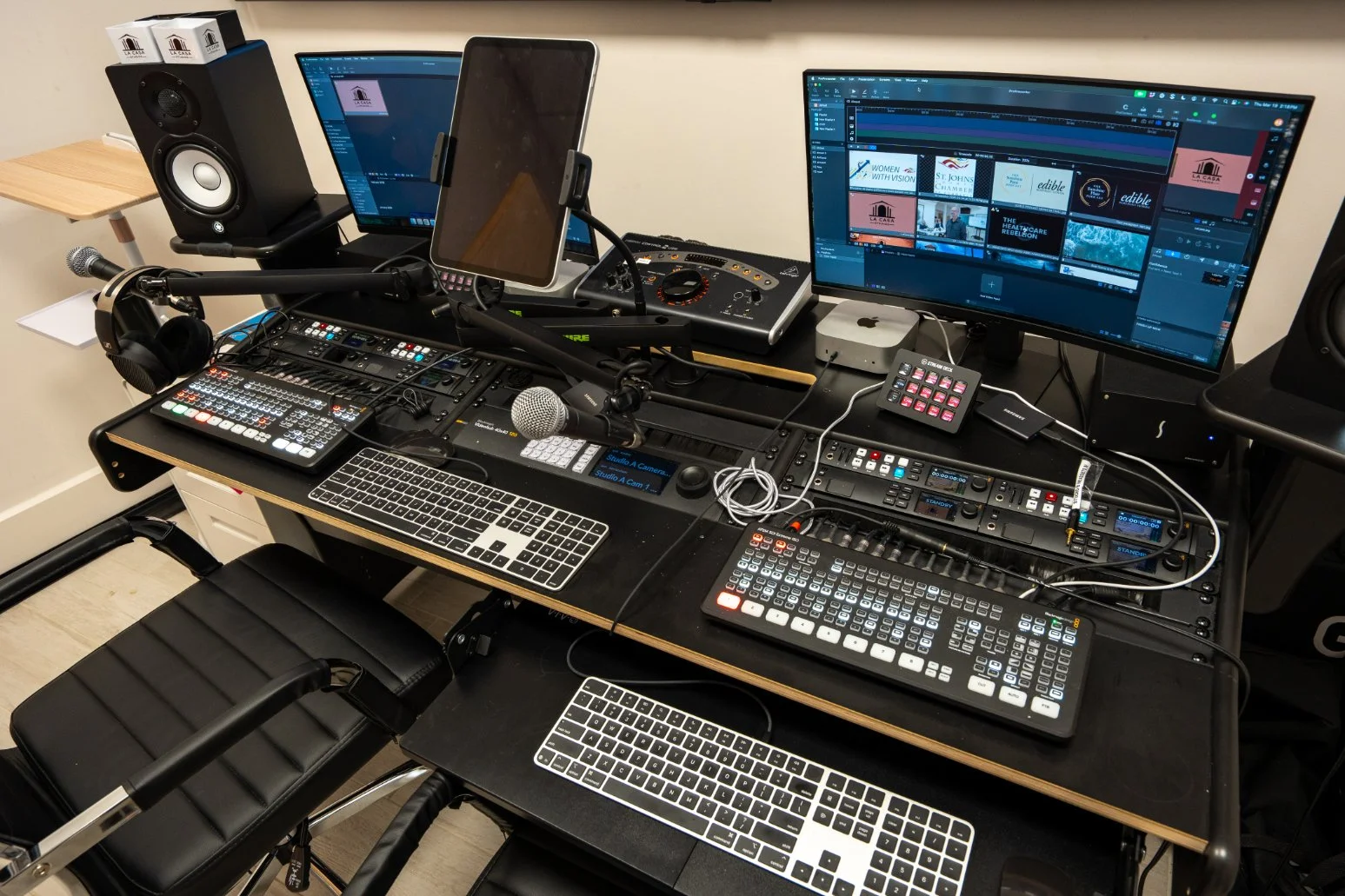 An audio recording studio workspace with two monitors displaying video editing software, a sound mixer, microphones, headphones, a small keyboard, and a chair at La Casa Studio in St. Augustine, FL