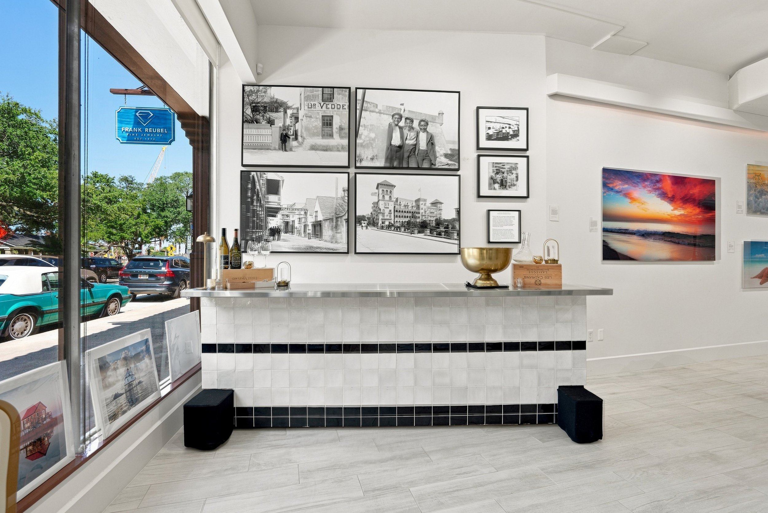 An art gallery interior featuring black-and-white photographs on the wall, with a wooden countertop displaying wine bottles and glassware, and with framed artwork on the lower part of the wall and a vibrant sunset photograph on the right wall.