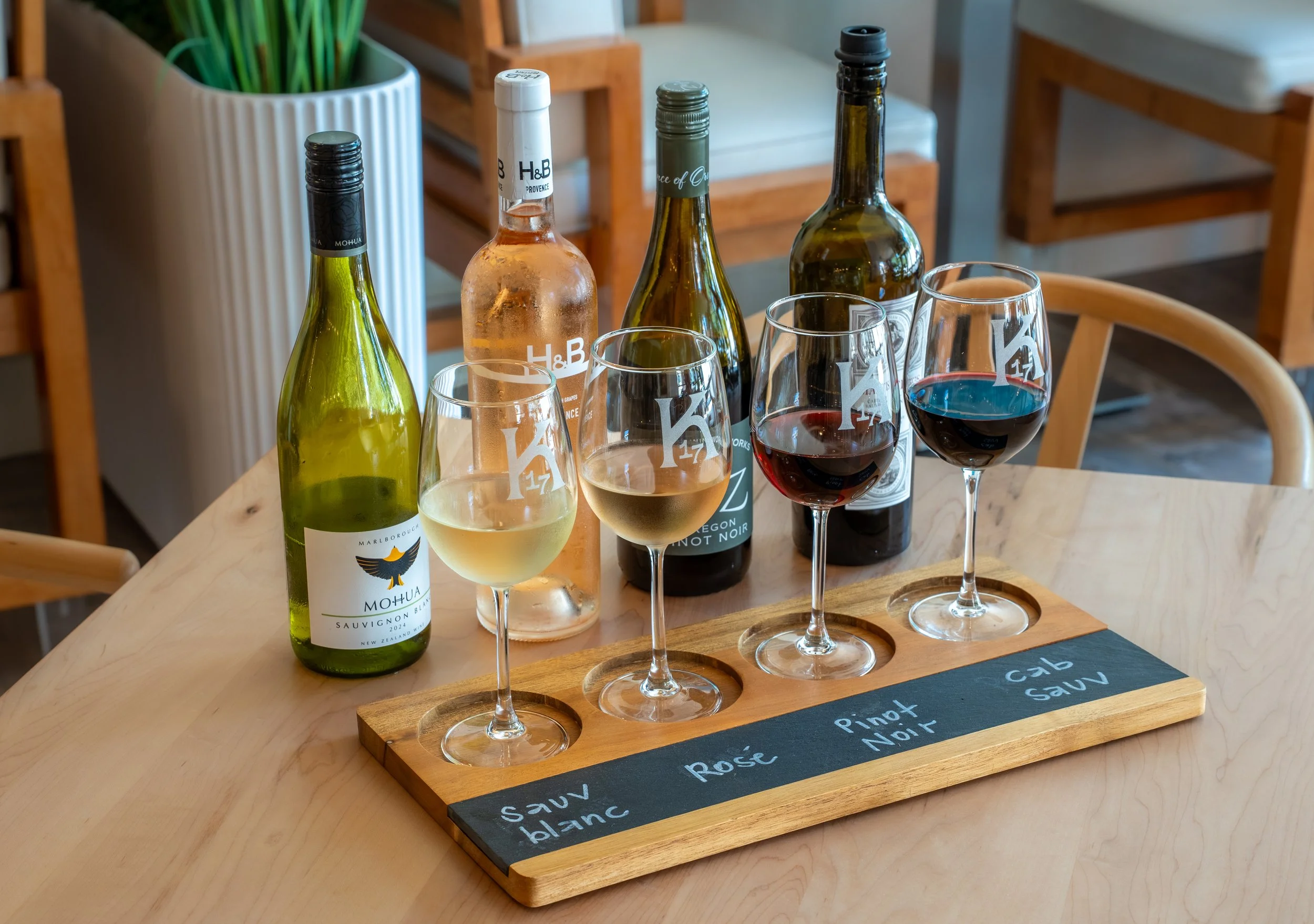 A wine tasting flight with four glasses of different wines and four bottles of wine on a wooden tray, set on a light wooden table in a cozy indoor setting.