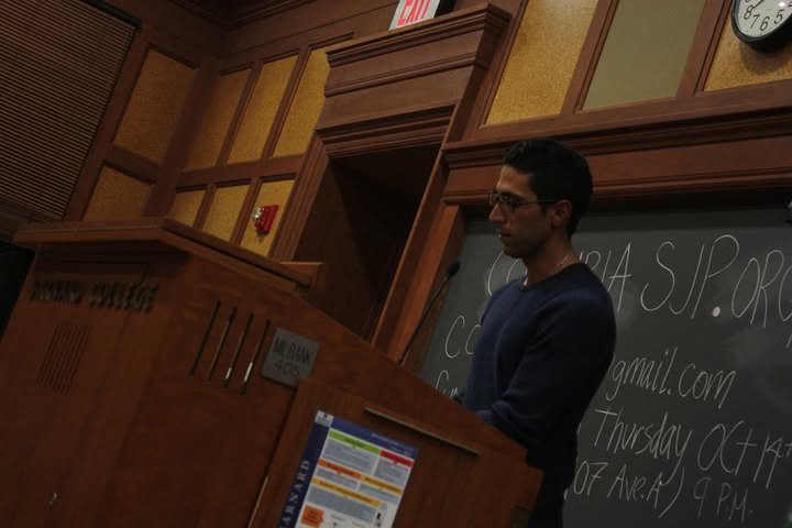 A man standing at a wooden podium in front of a blackboard with writing on it in a classroom or lecture room.