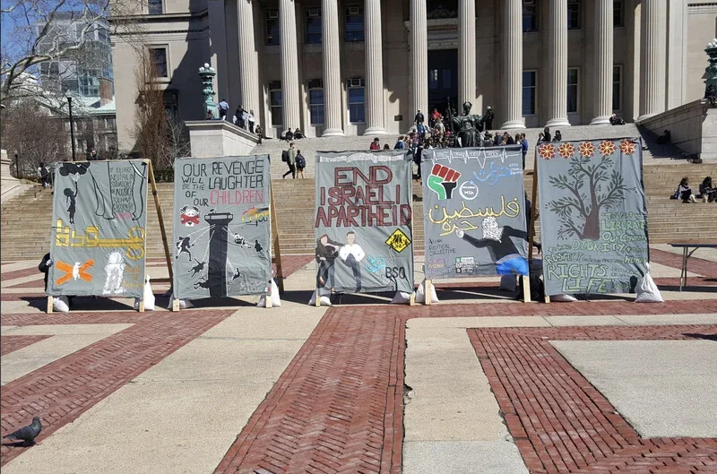 A series of protest posters on display outside a government building with steps, featuring messages about justice, ending Israeli apartheid, and supporting Palestinian rights, with some posters including artwork and slogans in English and Arabic.