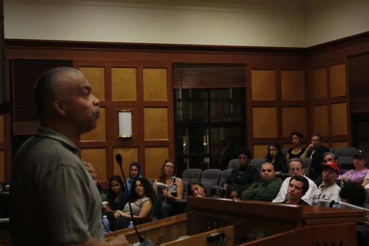 A man speaking at a podium in a courtroom or conference room with a seated audience of diverse men and women.
