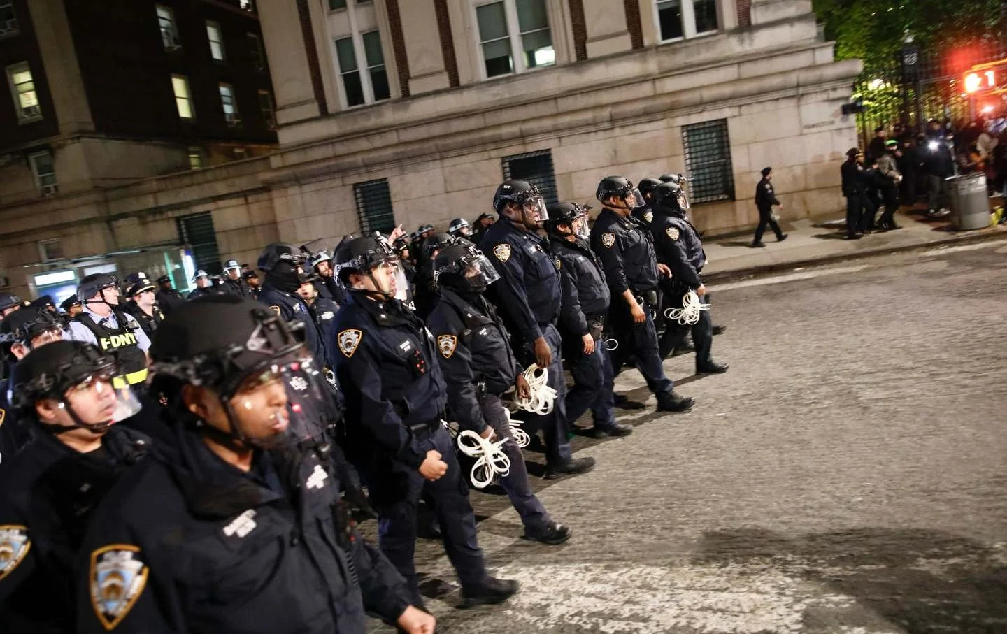 Line of police officers standing on a city street at night, wearing helmets and protective gear, holding ropes.
