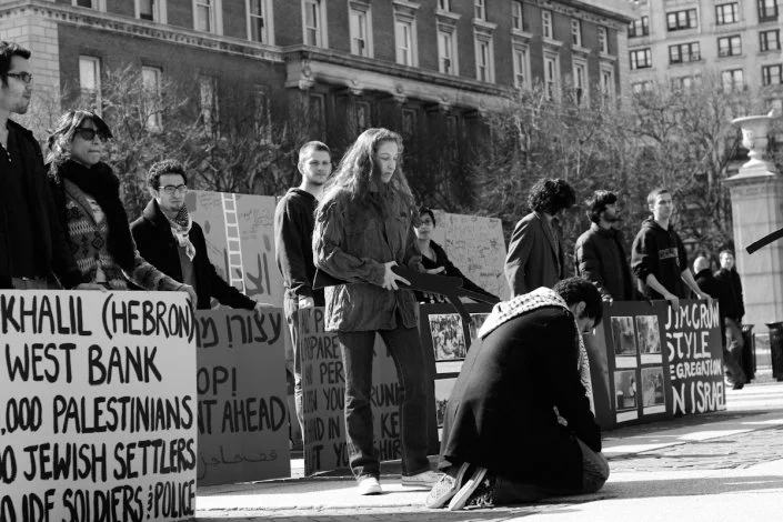 People participating in a protest or vigil, holding signs with messages about Palestine and other issues, some kneeling or standing outside in an urban area.