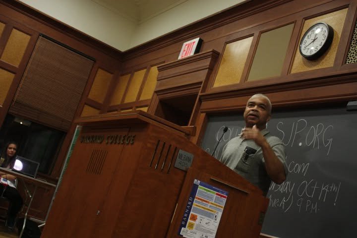 A man speaking at a wooden podium in a classroom or lecture hall, with a blackboard behind him and a clock on the wall showing 2:52. A woman is sitting at a desk with a laptop in the background.