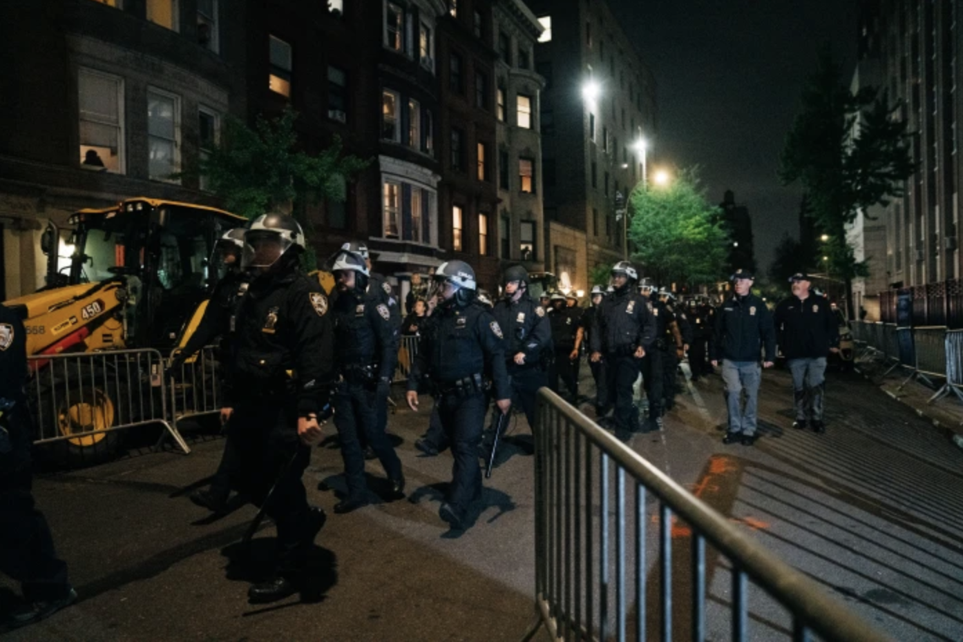 Nighttime street scene with police officers in uniform and riot gear, barricades, and buildings with lit windows.
