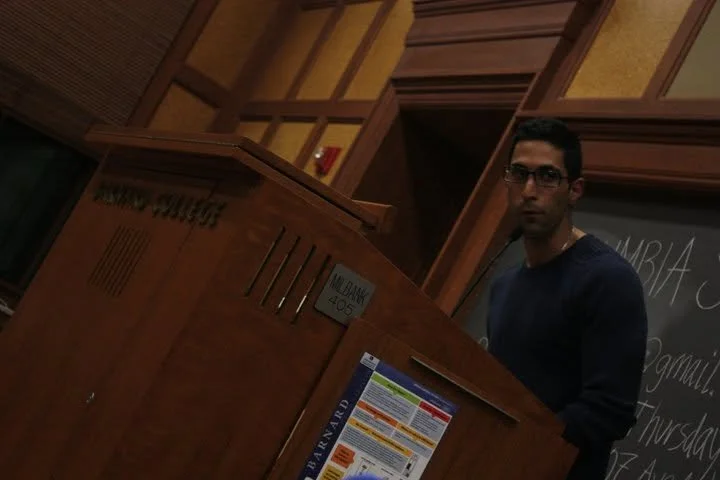A man with glasses and dark hair stands next to a wooden counter or podium, likely in a classroom or lecture hall, with a chalkboard in the background.