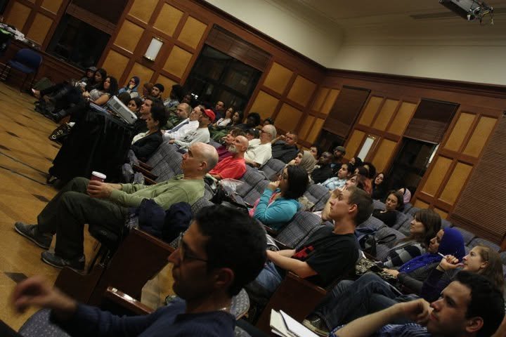 People attending a conference or seminar in a large, wood-paneled room, sitting in chairs and listening to a speaker at a podium.